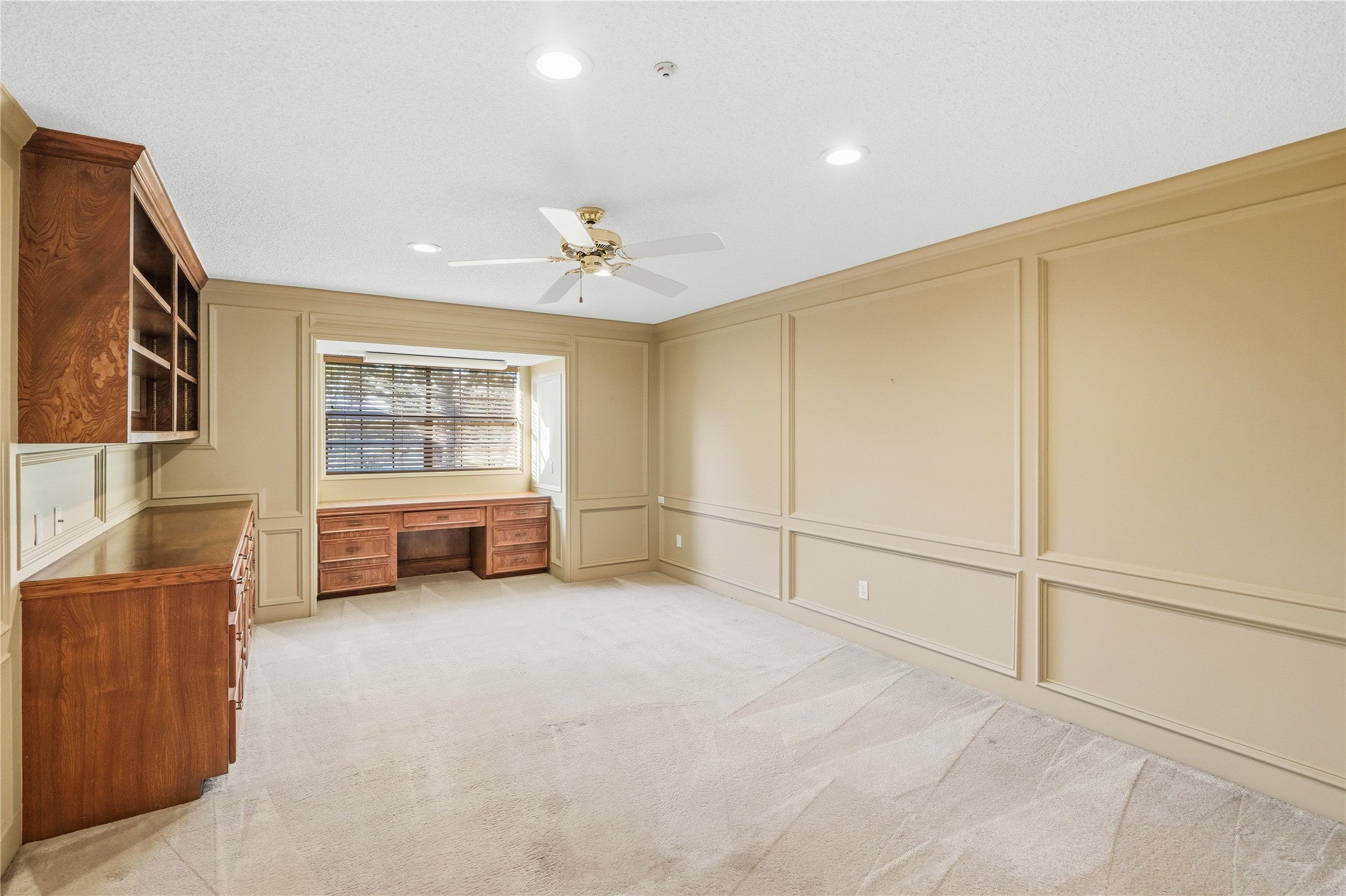 Guest room with wood paneling features built-in shelving and a desk. Natural light streams through a large window, complemented by recessed lighting and a ceiling fan for comfort.