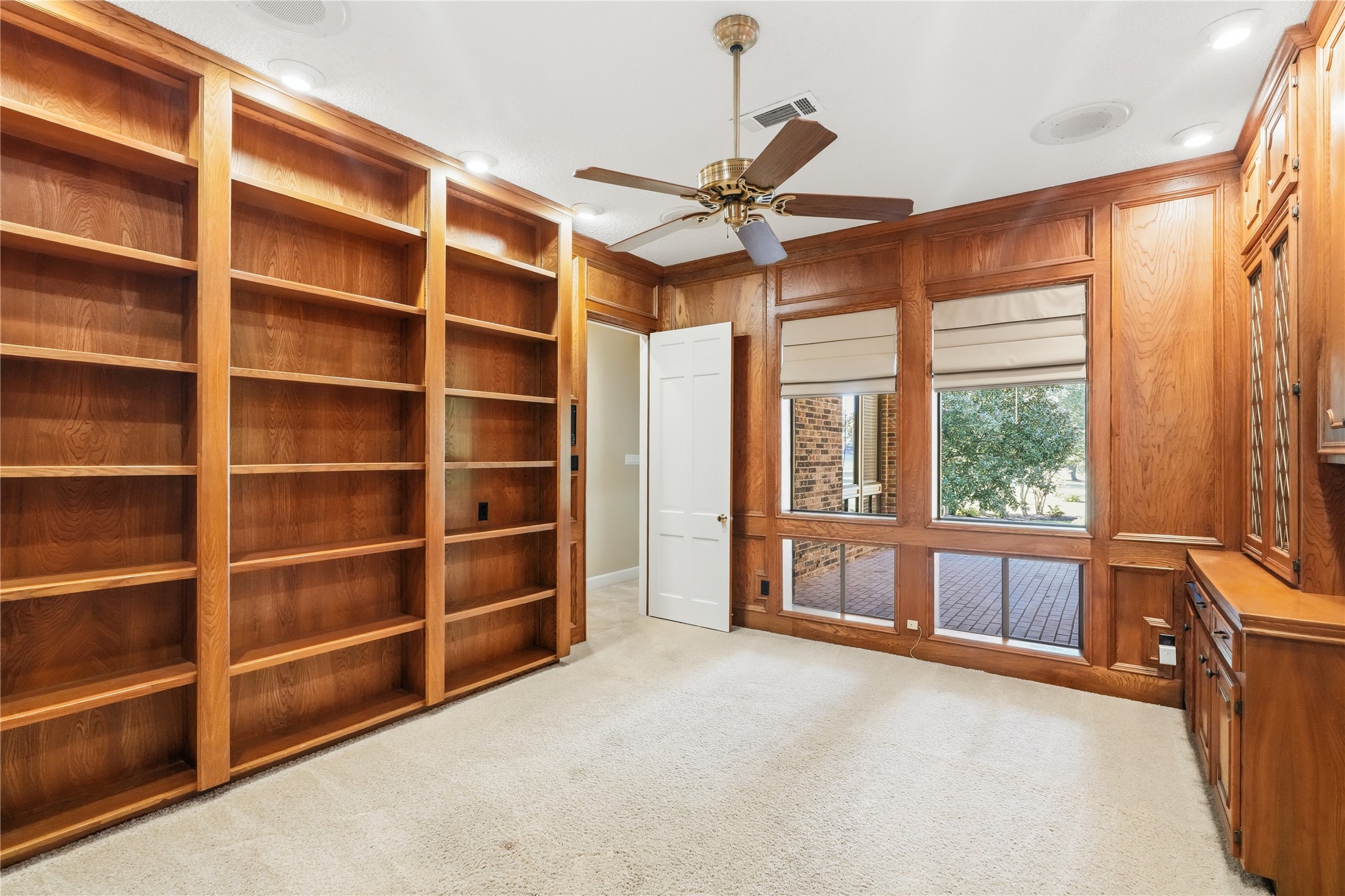 Home office featuring custom built-in wooden bookshelves, a ceiling fan, and large windows with views of the backyard, creating a warm and inviting workspace.