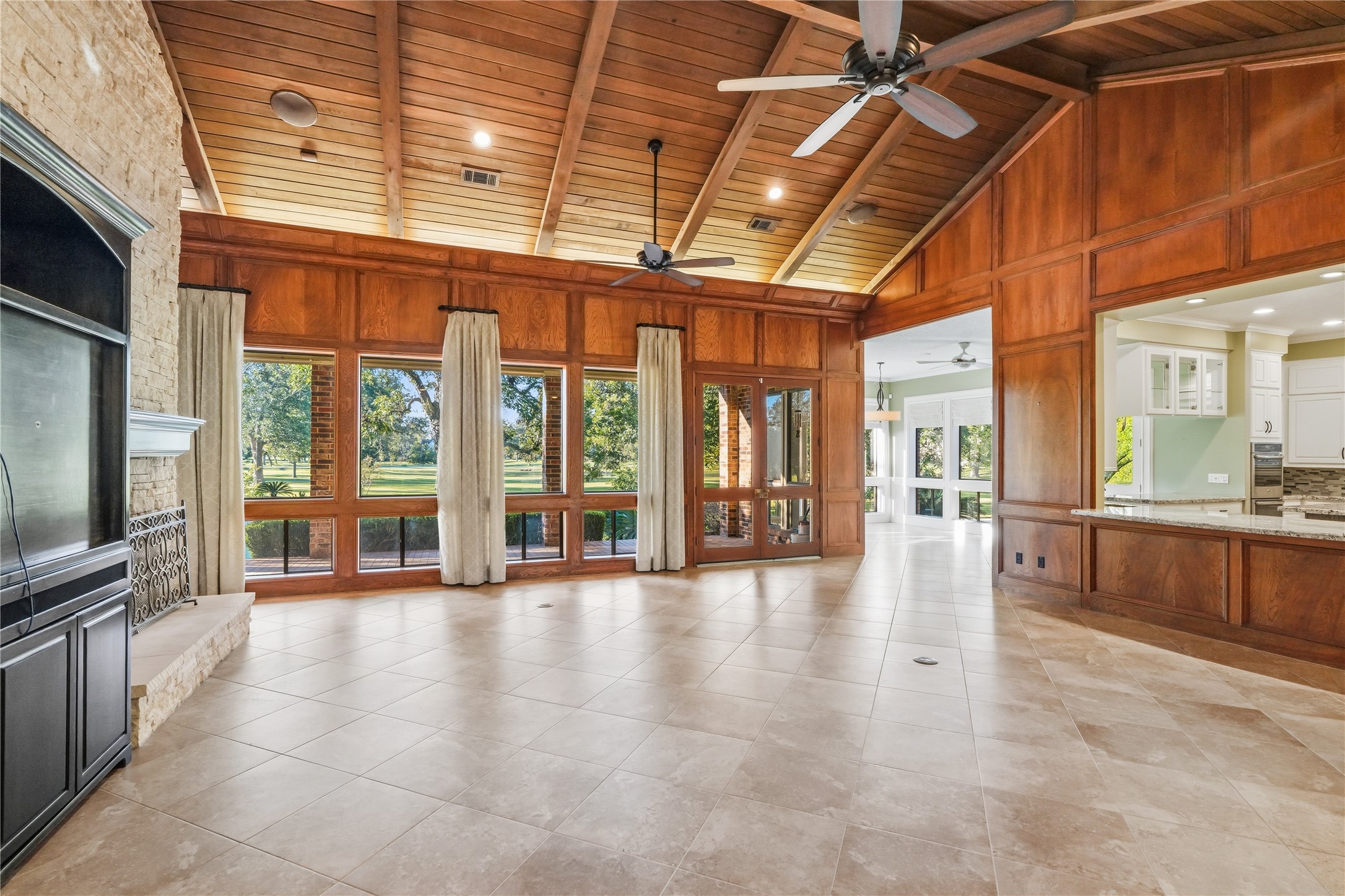 Spacious living area with vaulted wood ceilings, large windows offering stunning views, and a cozy stone fireplace, seamlessly connected to the bright kitchen.