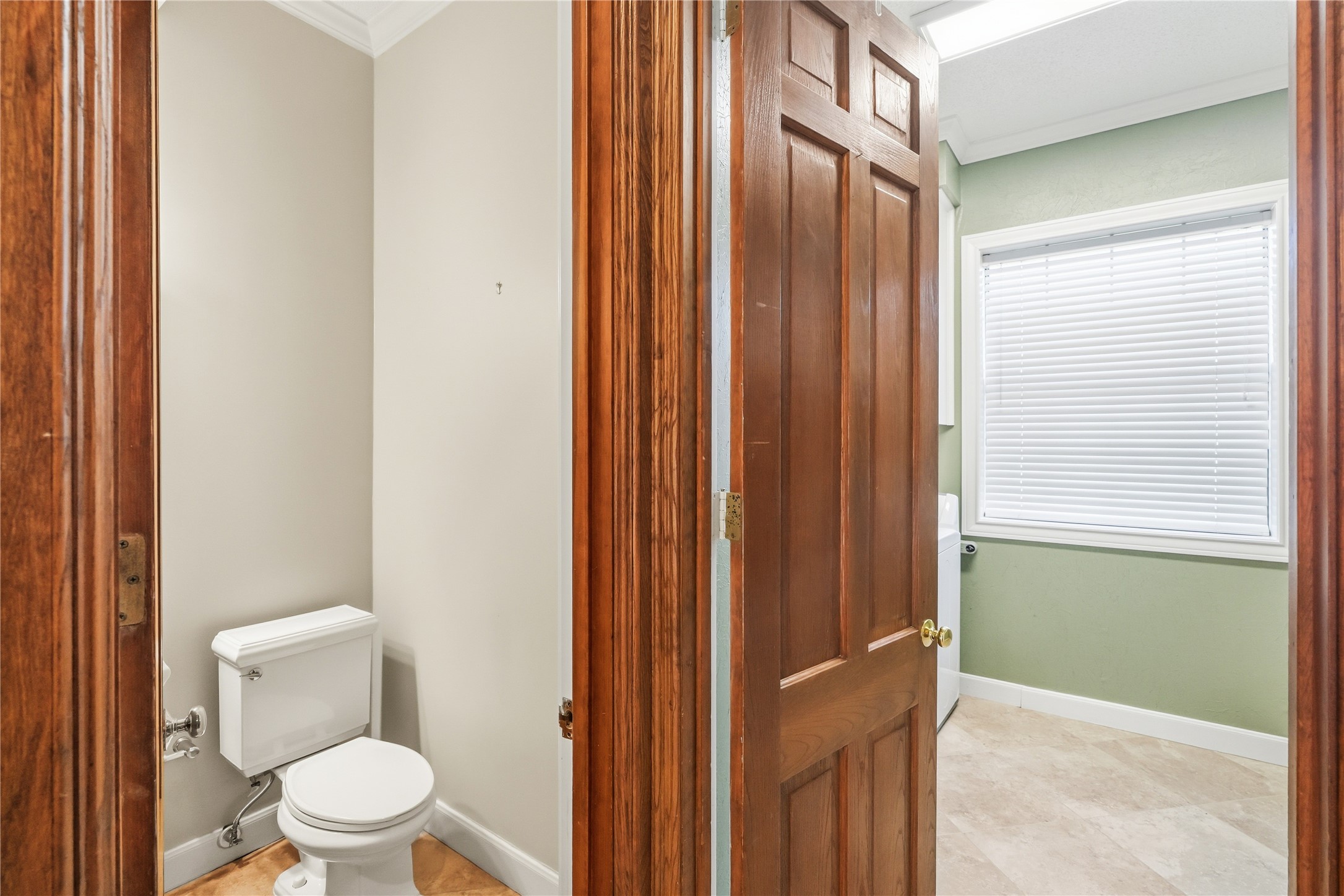 Powder room with elegant wooden door, adjacent to a bright laundry room featuring a window with blinds, offering privacy and natural light.