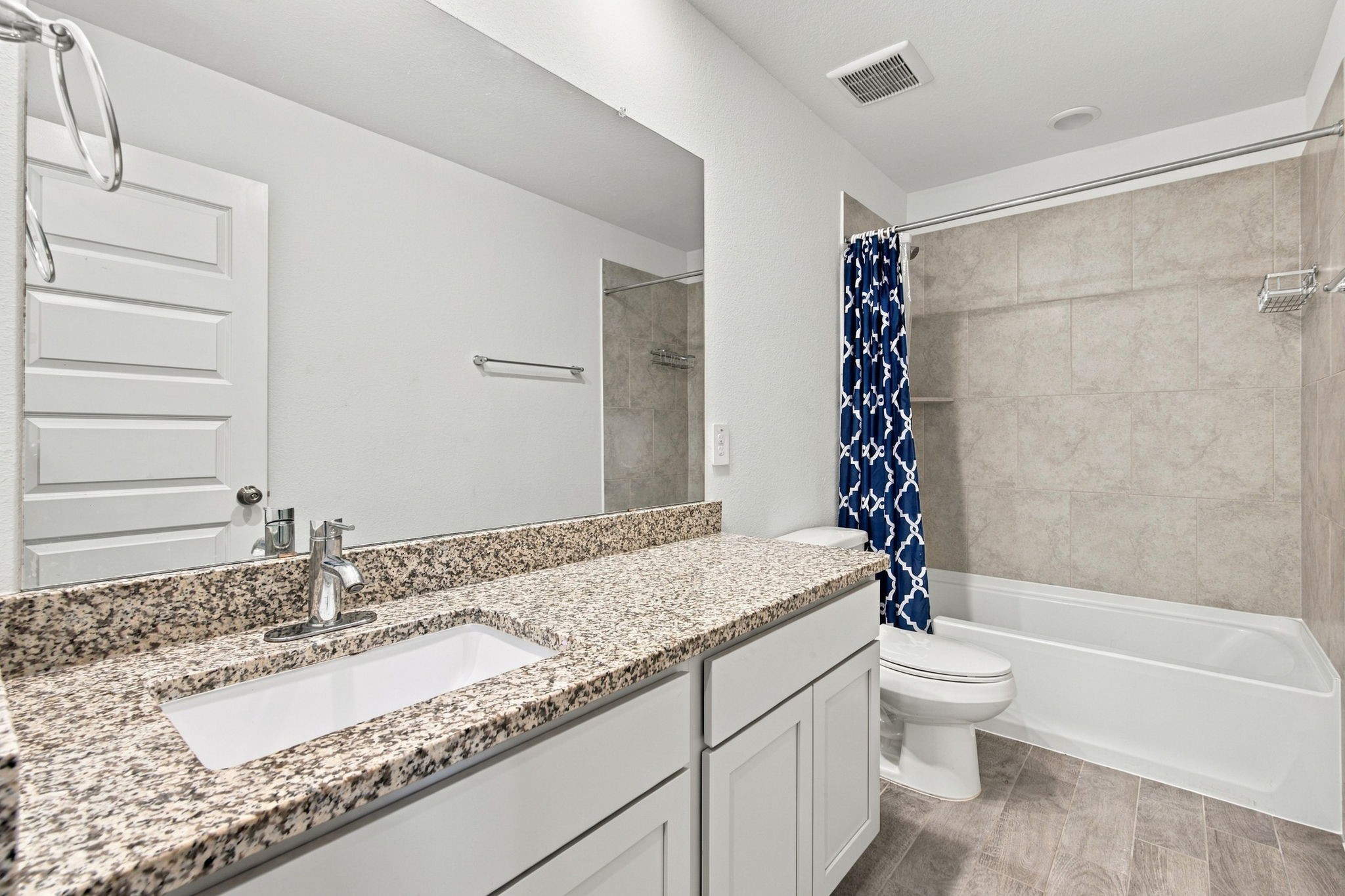 This bright and spacious full bathroom features a clean, modern design with crisp white cabinetry, beautiful speckled granite countertops, and a generous vanity offering ample counter space and storage. The shower and bathtub combination features large-format neutral-toned tile surrounds, creating a stylish and low-maintenance bathing space.