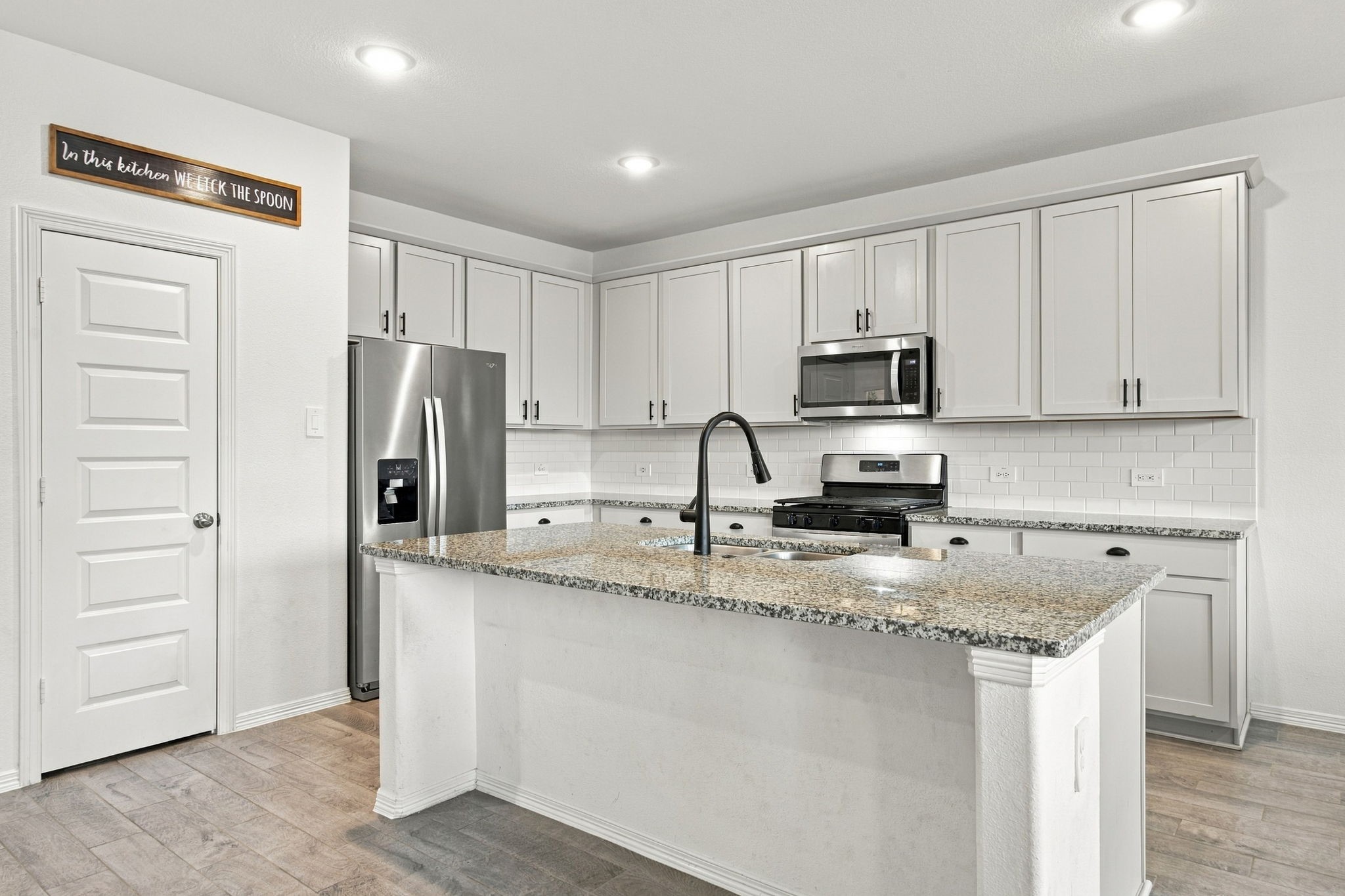 This bright and spacious kitchen features a modern, functional layout, beautiful speckled granite countertops, and a classic white subway tile backsplash. A generously sized center island offers ample preparation space, an integrated undermount sink, and a sleek black gooseneck faucet. Stainless steel appliances, including a French-door refrigerator, gas range, and built-in microwave, complement the kitchen's clean, contemporary finishes.