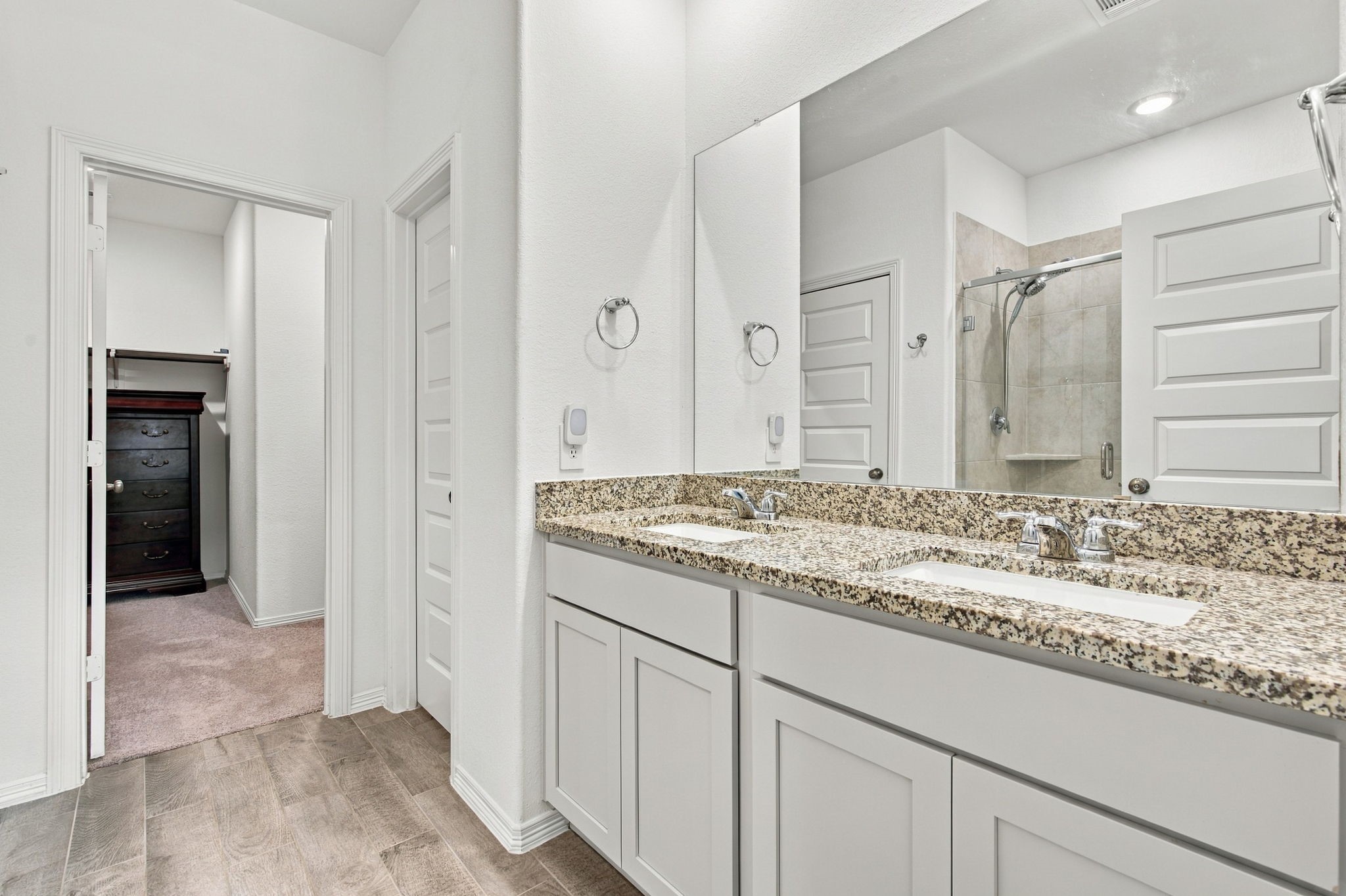 This bright and spacious primary bathroom features a beautiful double-sink vanity with speckled granite countertops, crisp white cabinetry, and chrome fixtures. A large wall-mounted mirror enhances the open feel of the space, while recessed ceiling lighting provides excellent illumination.