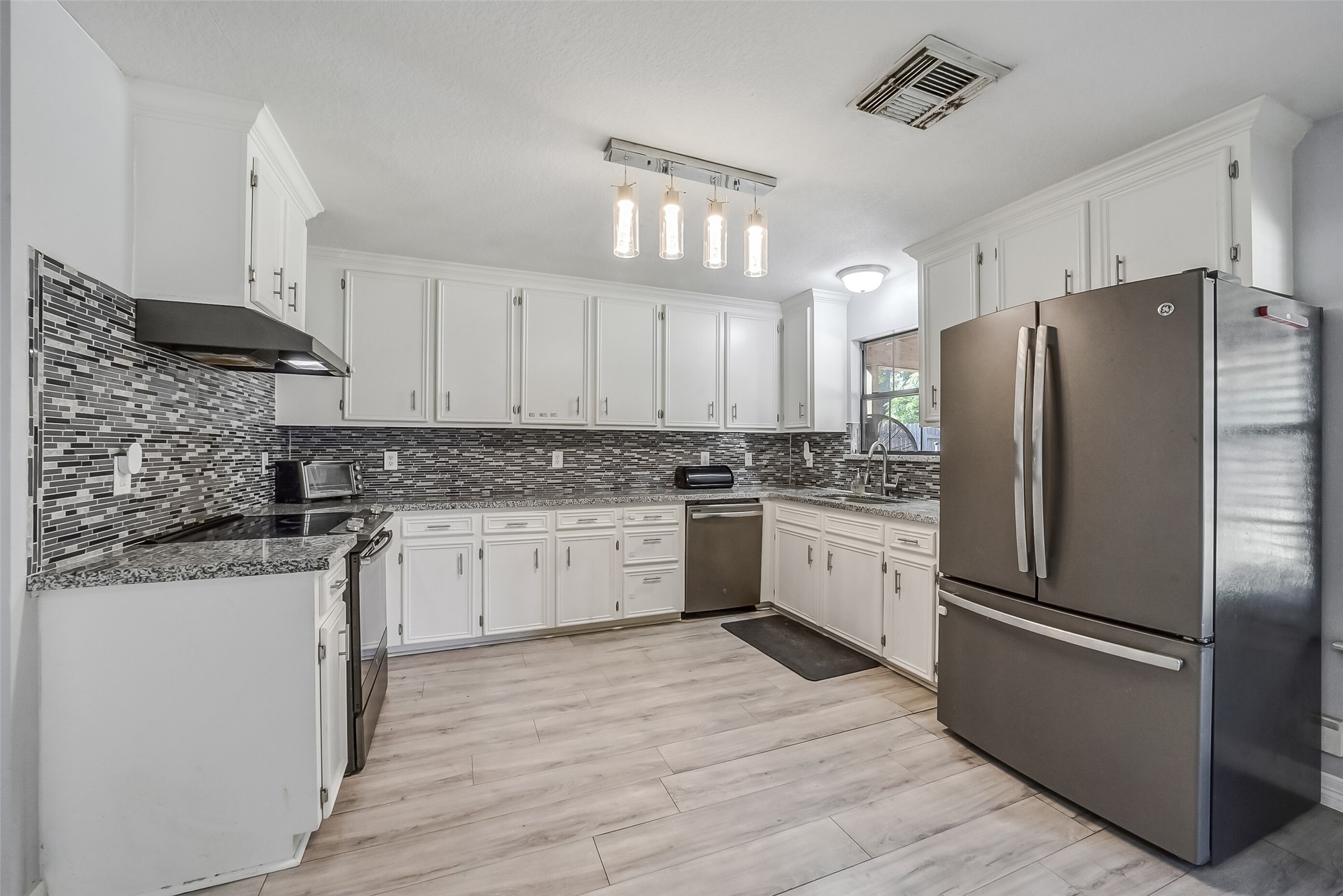 This bright kitchen features abundant white cabinetry, granite countertops, and a striking mosaic tile backsplash. Stainless steel appliances, contemporary lighting, and wood-look flooring complement the space, while a window above the sink welcomes natural light.