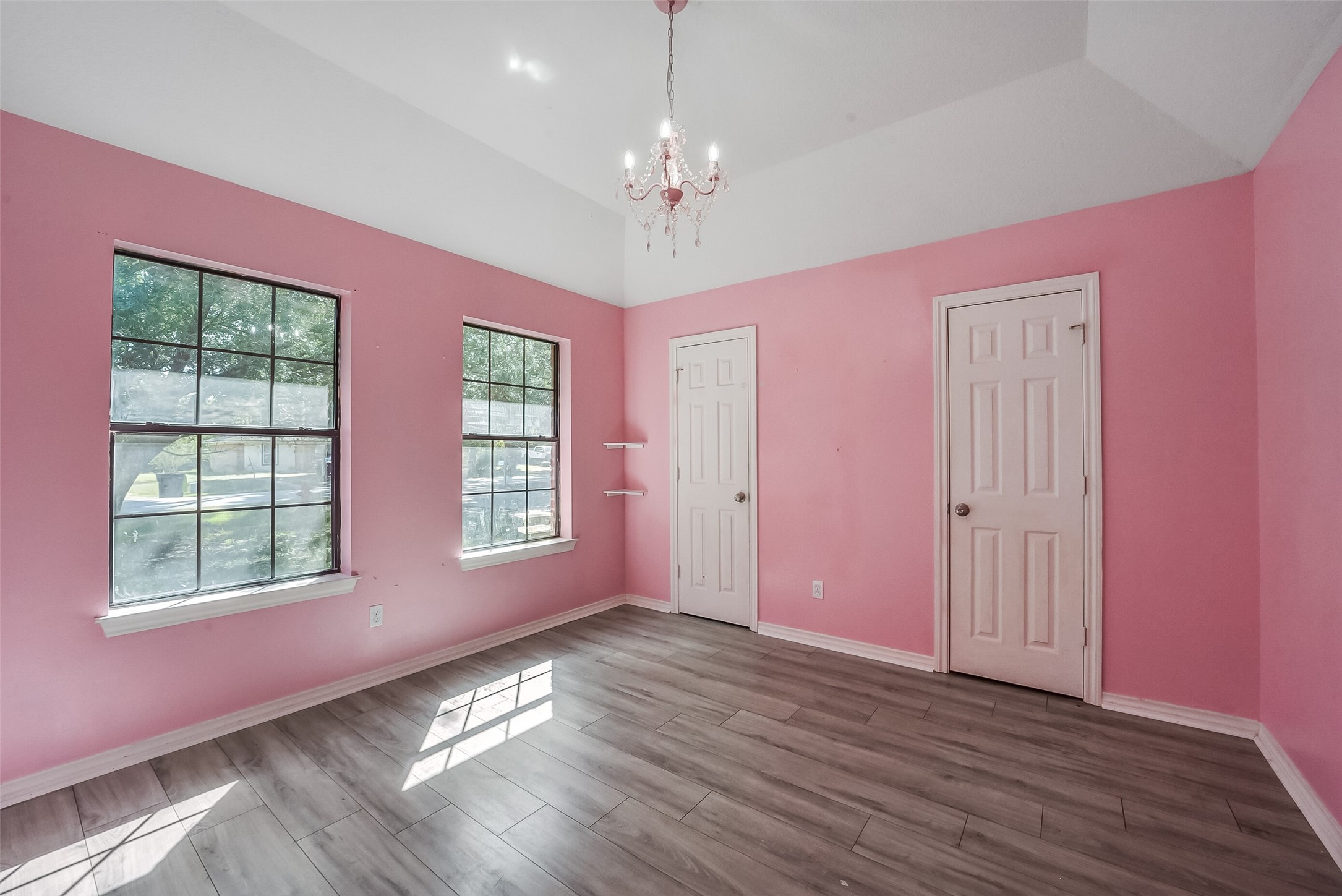 This charming secondary bedroom features high ceilings, a decorative chandelier, and two large windows that fill the space with natural light. Pink walls, crisp white trim, and wood-look flooring create a bright, inviting atmosphere, while corner shelves offer a thoughtful decorative touch.