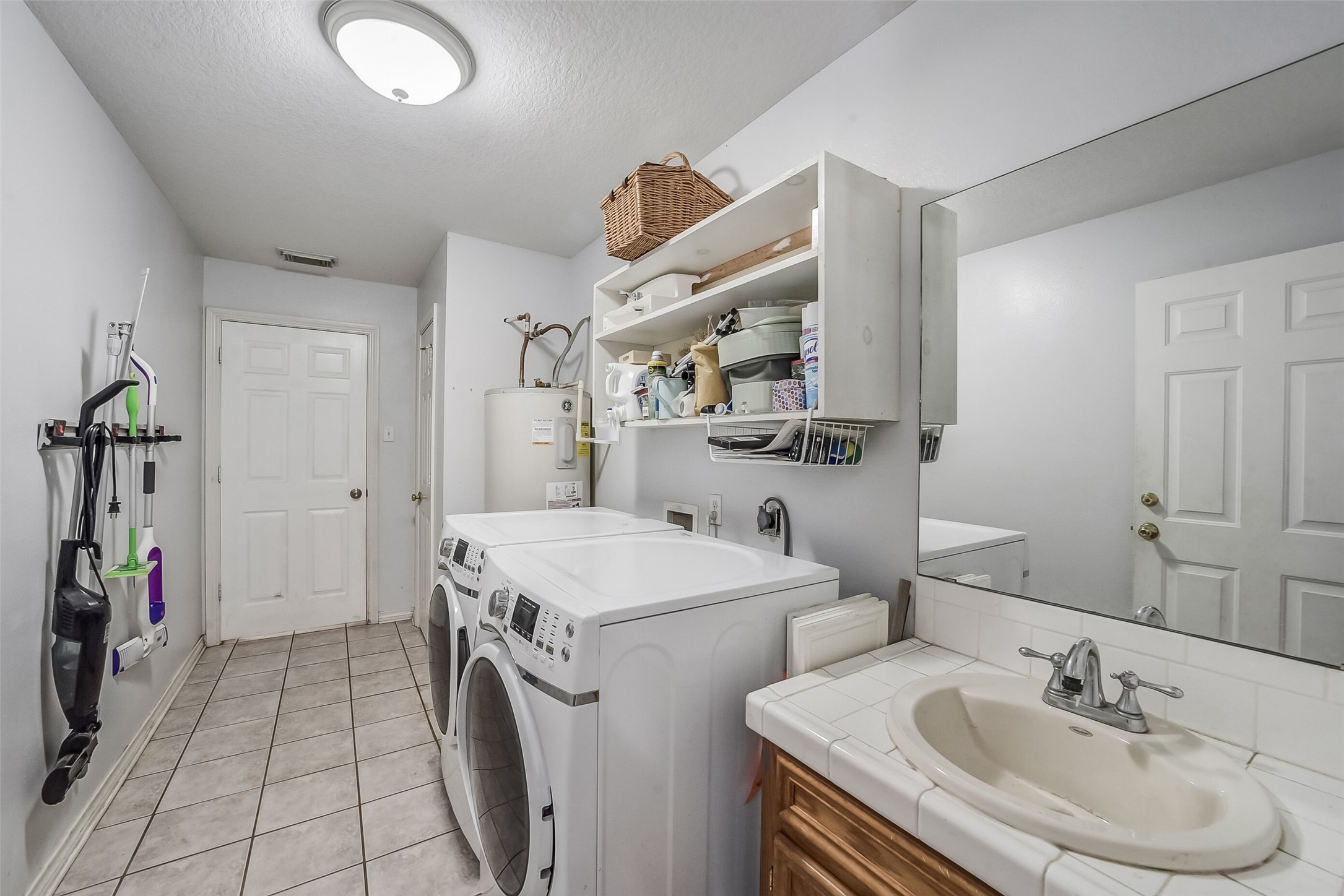 This functional laundry room features tile flooring, overhead shelving for convenient storage, and space for a full-size washer and dryer. A sink with a tiled countertop and lower cabinetry provides added convenience for everyday tasks.