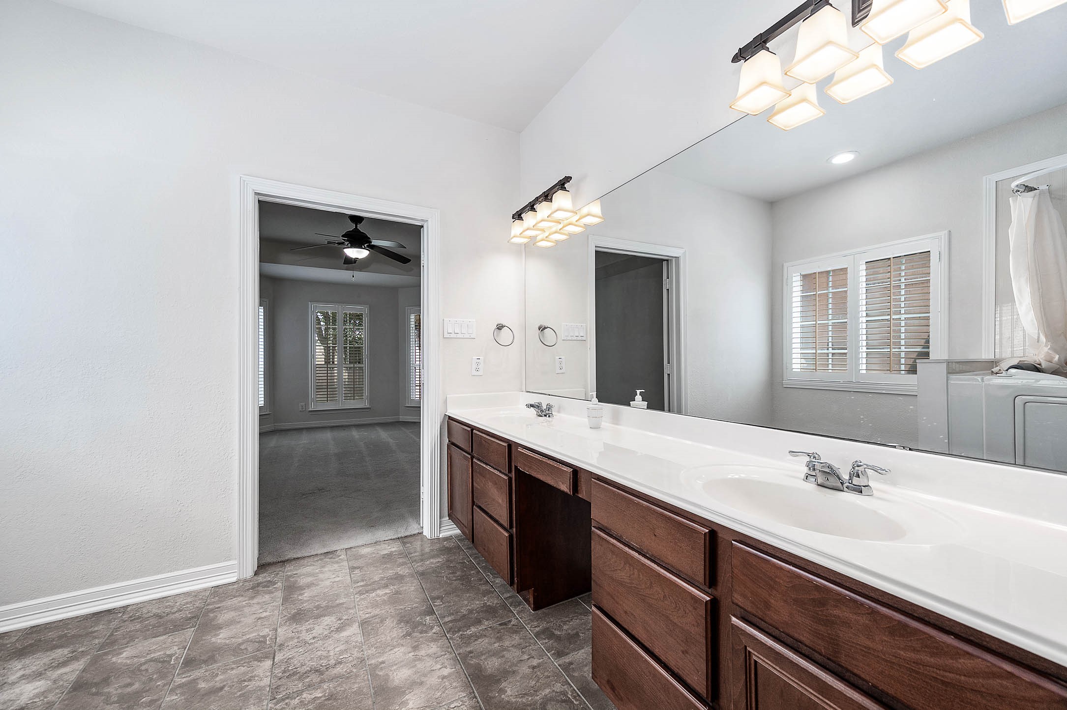 Spacious bathroom featuring a dual-sink vanity with ample counter space and elegant lighting, leading into a cozy carpeted room with natural light from shuttered windows.