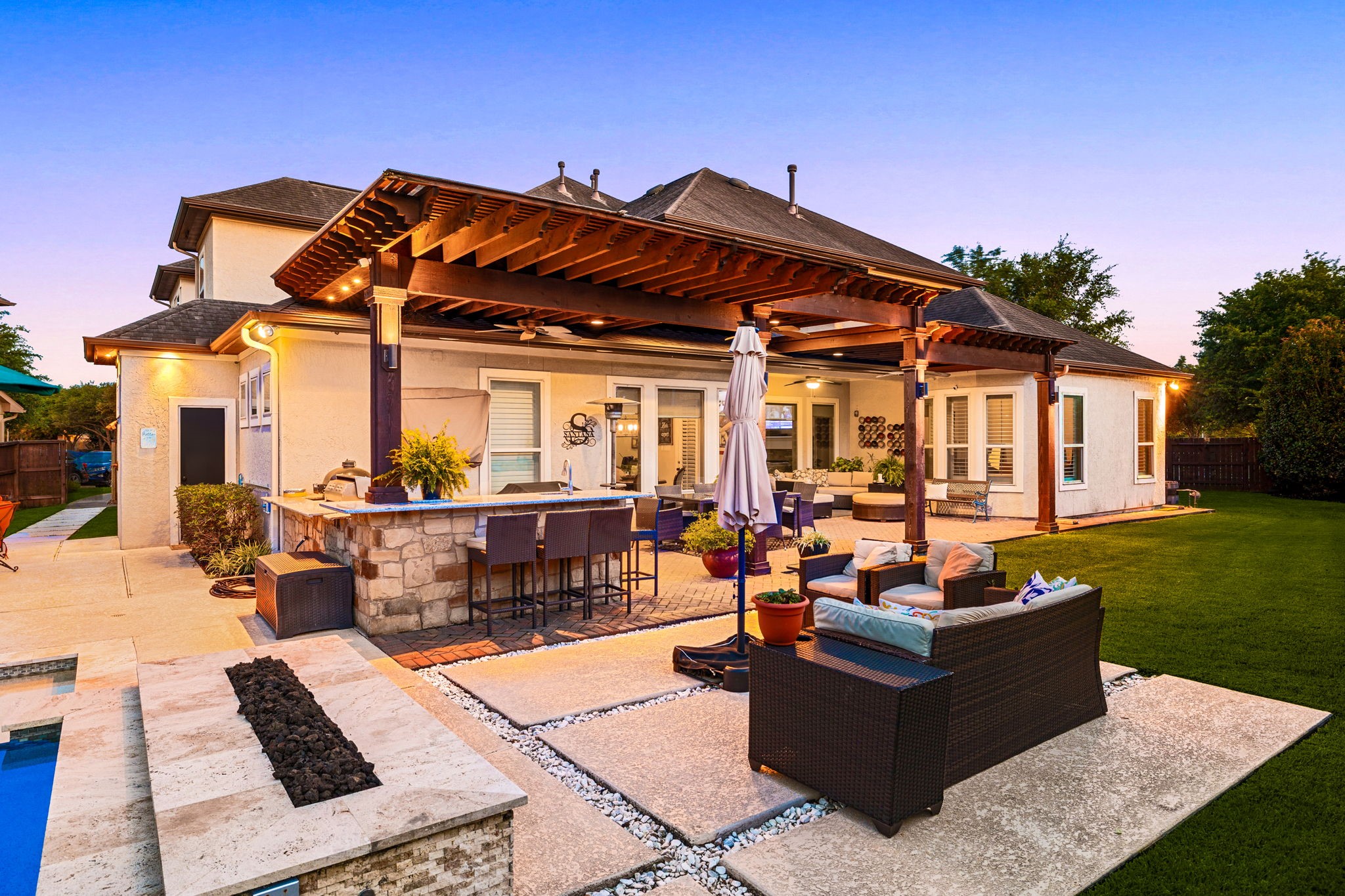 Backyard goals at golden hour! This breathtaking outdoor living space features a grand wood pergola with recessed lighting, a stacked stone outdoor kitchen and bar, built-in fire feature, travertine and brick paver patio, and lush green lawn, the ultimate entertainer's dream under the Texas sky!