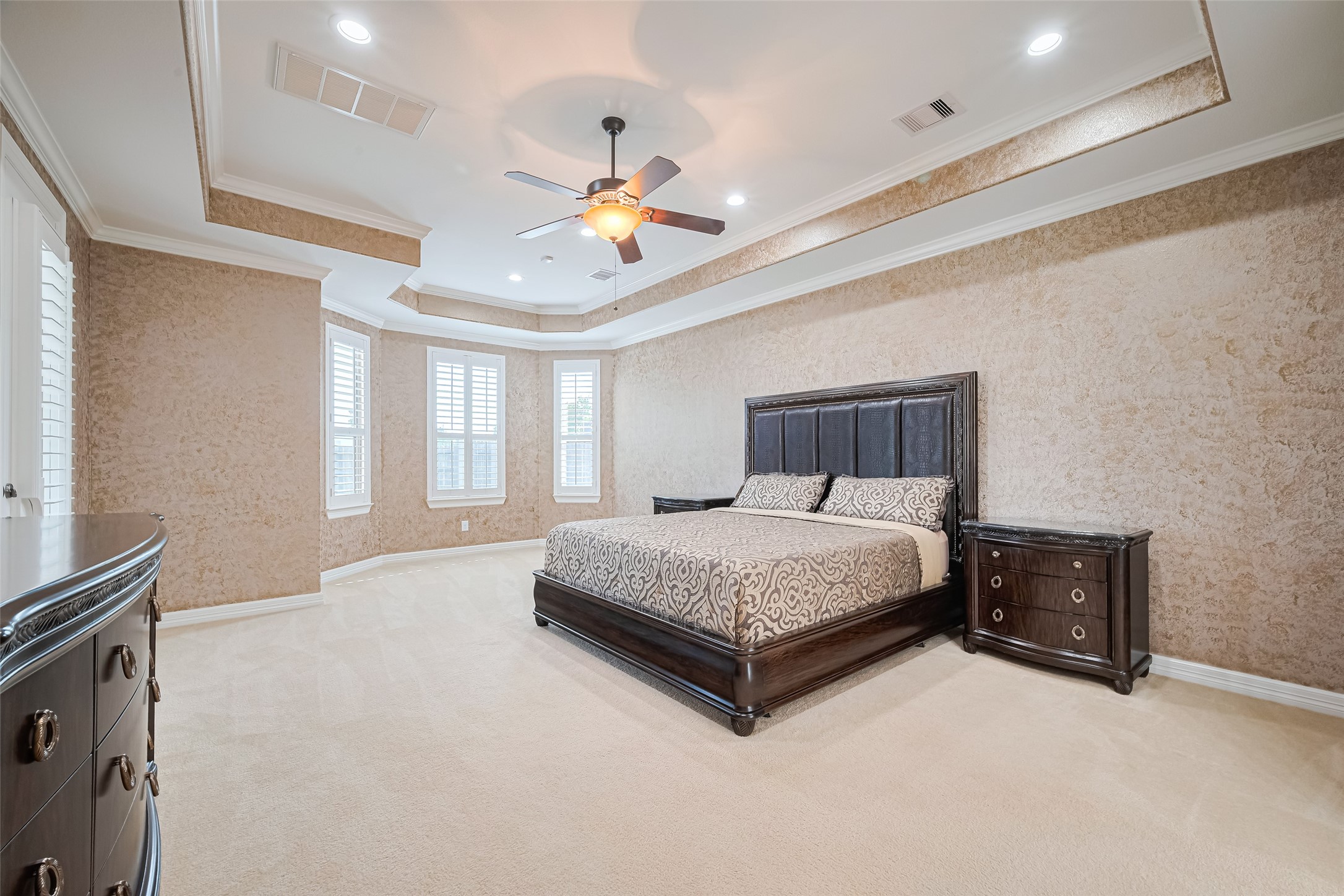 Expansive primary suite enhanced by an intricate tray ceiling, textured finishes, and a sunlit bay of plantation-shuttered windows.