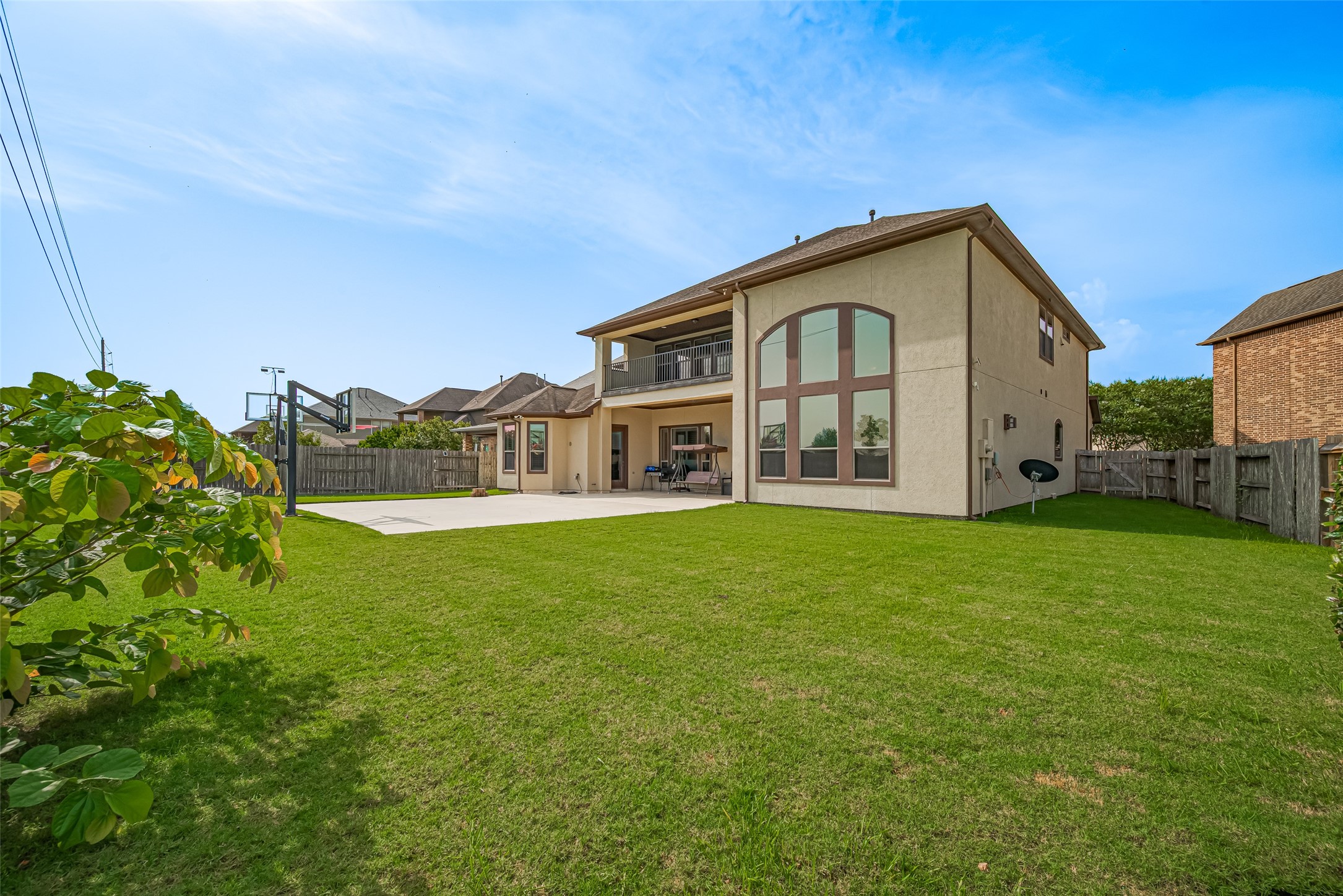 Expansive fenced backyard with a lush lawn, basketball court, covered patio, upper balcony, and striking rear elevation with dramatic oversized windows.