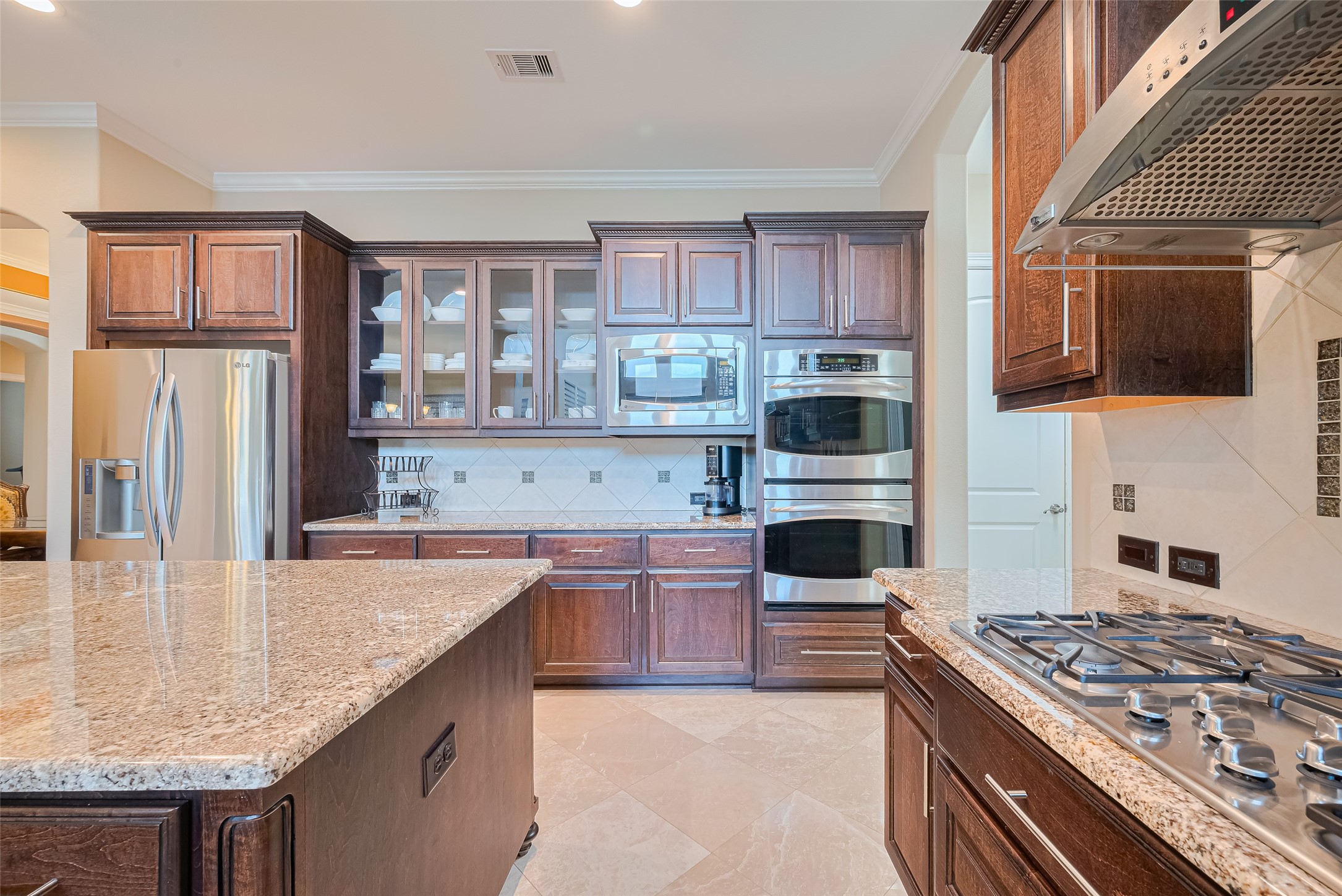 Detailed culinary view showcasing the five-burner gas cooktop, vent hood, stacked ovens, glass-front cabinets, and stainless steel refrigerator.