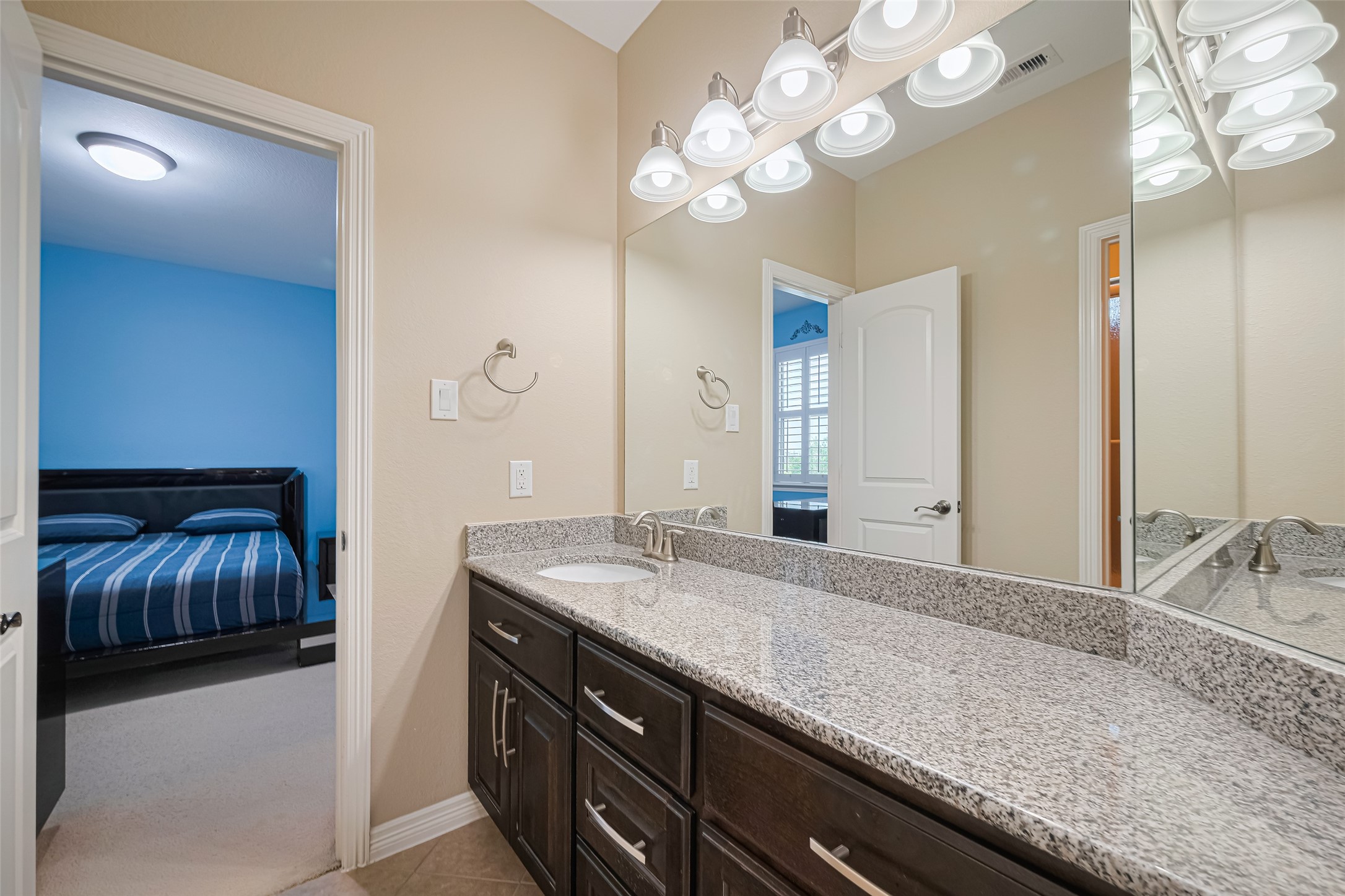 Bathroom featuring a spacious granite vanity, dark cabinetry, expansive mirror, and convenient access from the adjoining bedroom.