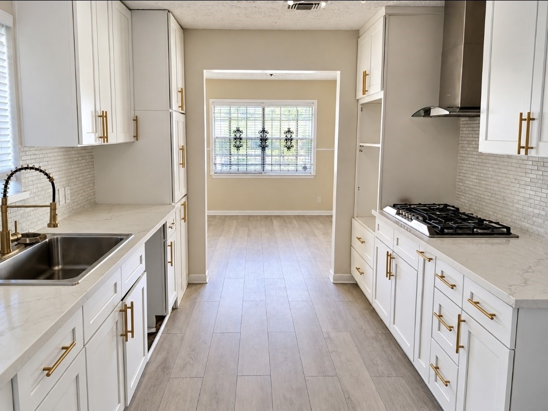 Bright and modern kitchen featuring sleek white cabinetry with gold hardware, a stylish backsplash, and stainless steel appliances, leading to a cozy dining area with natural light.