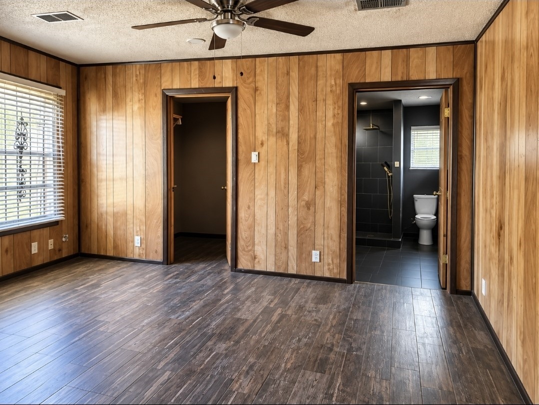 Spacious room with rich wood paneling, hardwood floors, and a ceiling fan, featuring an ensuite bathroom with sleek black tile and modern fixtures.