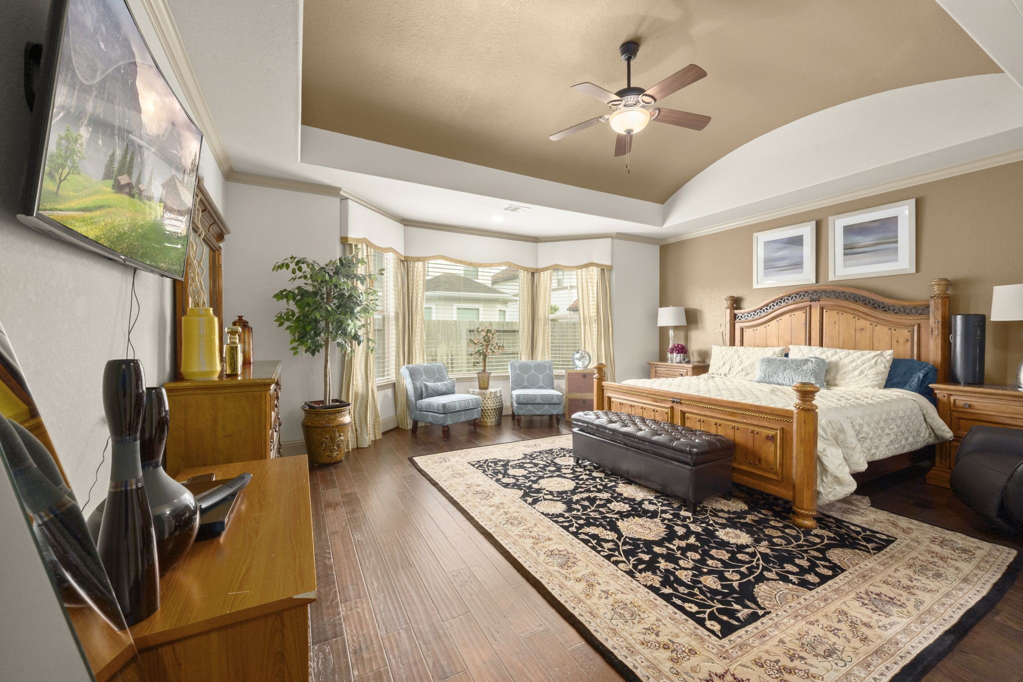 This spacious primary bedroom features elegant hardwood floors, a vaulted ceiling, and ample natural light from large windows.