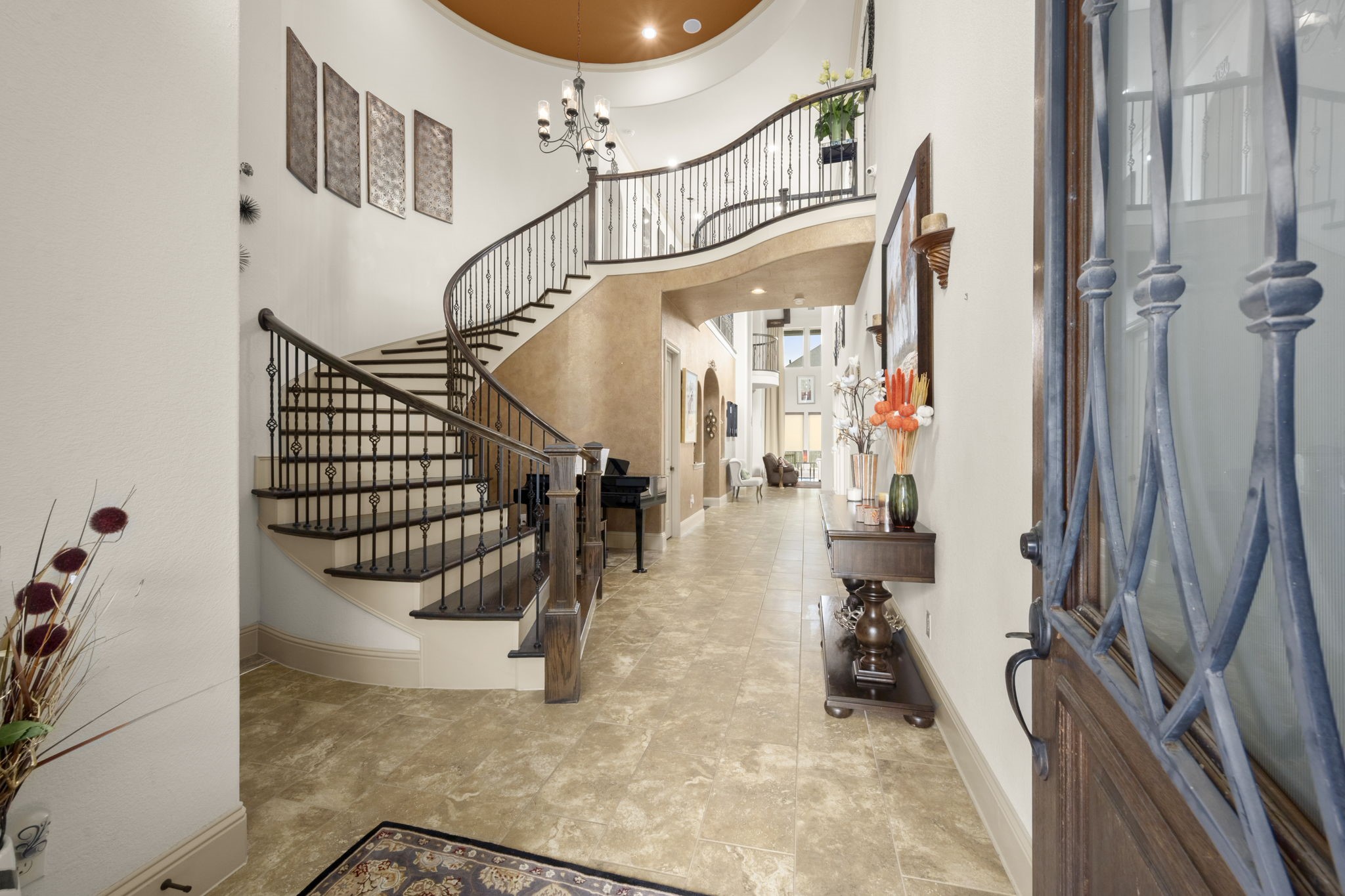 The home features an elegant foyer with a grand, curved staircase and wrought iron railing. This space also features high ceilings and a chandelier that leads into a well-lit hallway with tile flooring.