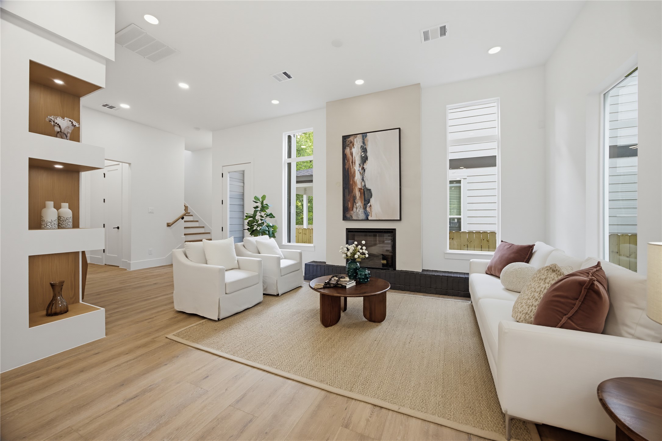 Modern living room with high ceilings, featuring a cozy fireplace and abundant natural light from tall windows. The open layout includes elegant built-in shelving and warm wood flooring, creating a welcoming ambiance.