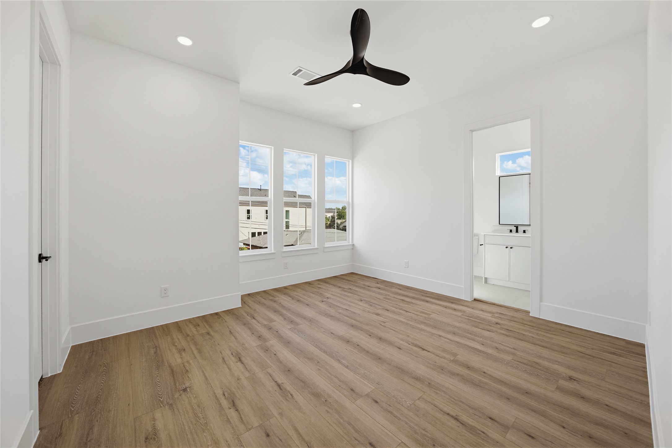 Bright and airy 3rd bedroom featuring large windows, sleek ceiling fan, and elegant wood flooring, leading to an en-suite bathroom.