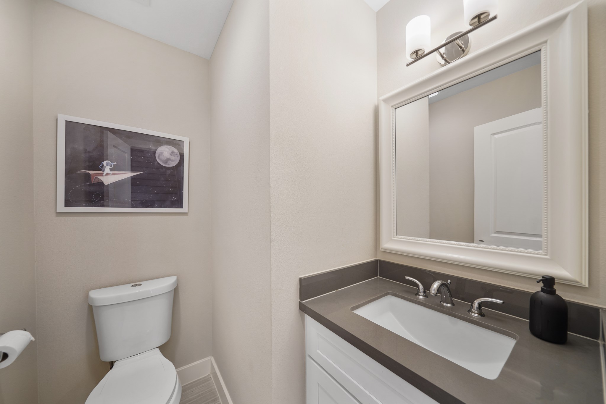Convenient for guests, the half bath includes a painted Shaker-style cabinet with a quartz counter top and integrated back splash accompanied by a framed mirror with wall mounted lighting above & tile flooring.