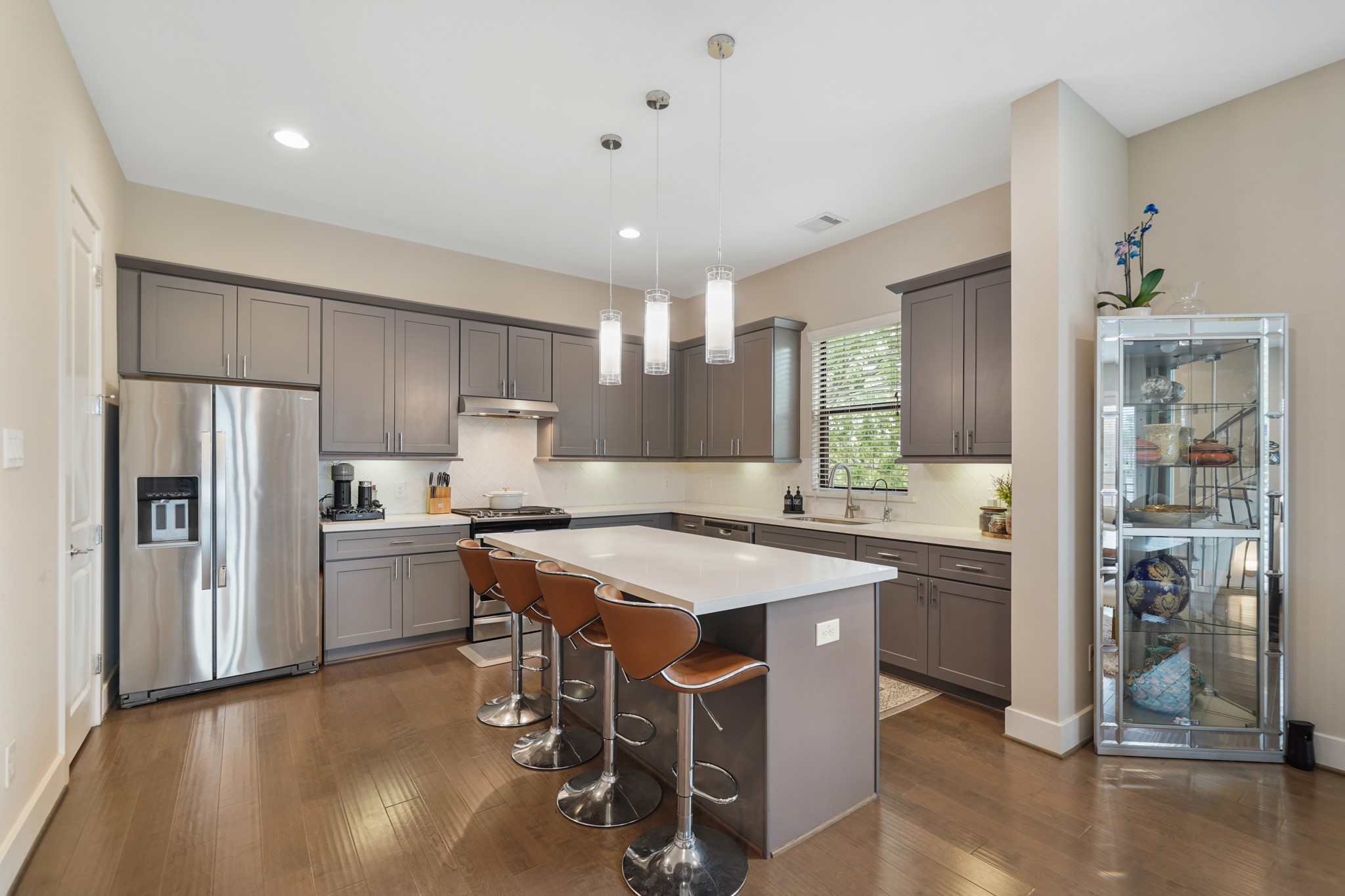 The kitchen sits on the back of the home with a high ceiling, 42" painted Shaker-style cabinets capped with crown moulding, quartz counter tops highlighted by a herringbone patterned and subway tiled back splash with under cabinet lighting & hardwood floors.