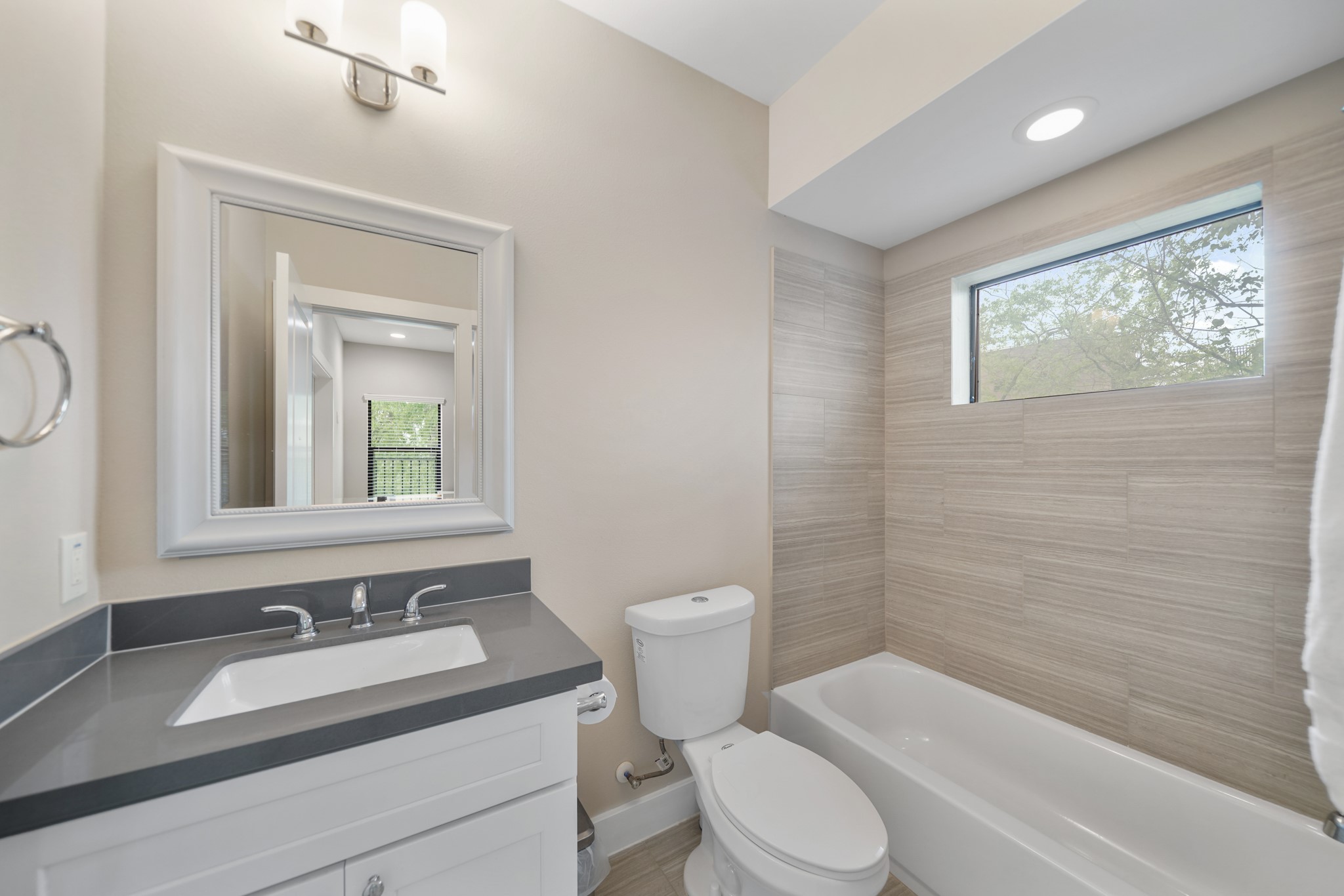 Serving as the home's third full bath, this bath includes a painted Shaker-style cabinet supporting a quartz counter with integrated back splash, framed mirror with lighting above, a ceramic tile surrounded combination tub/shower with window above & tile flooring.