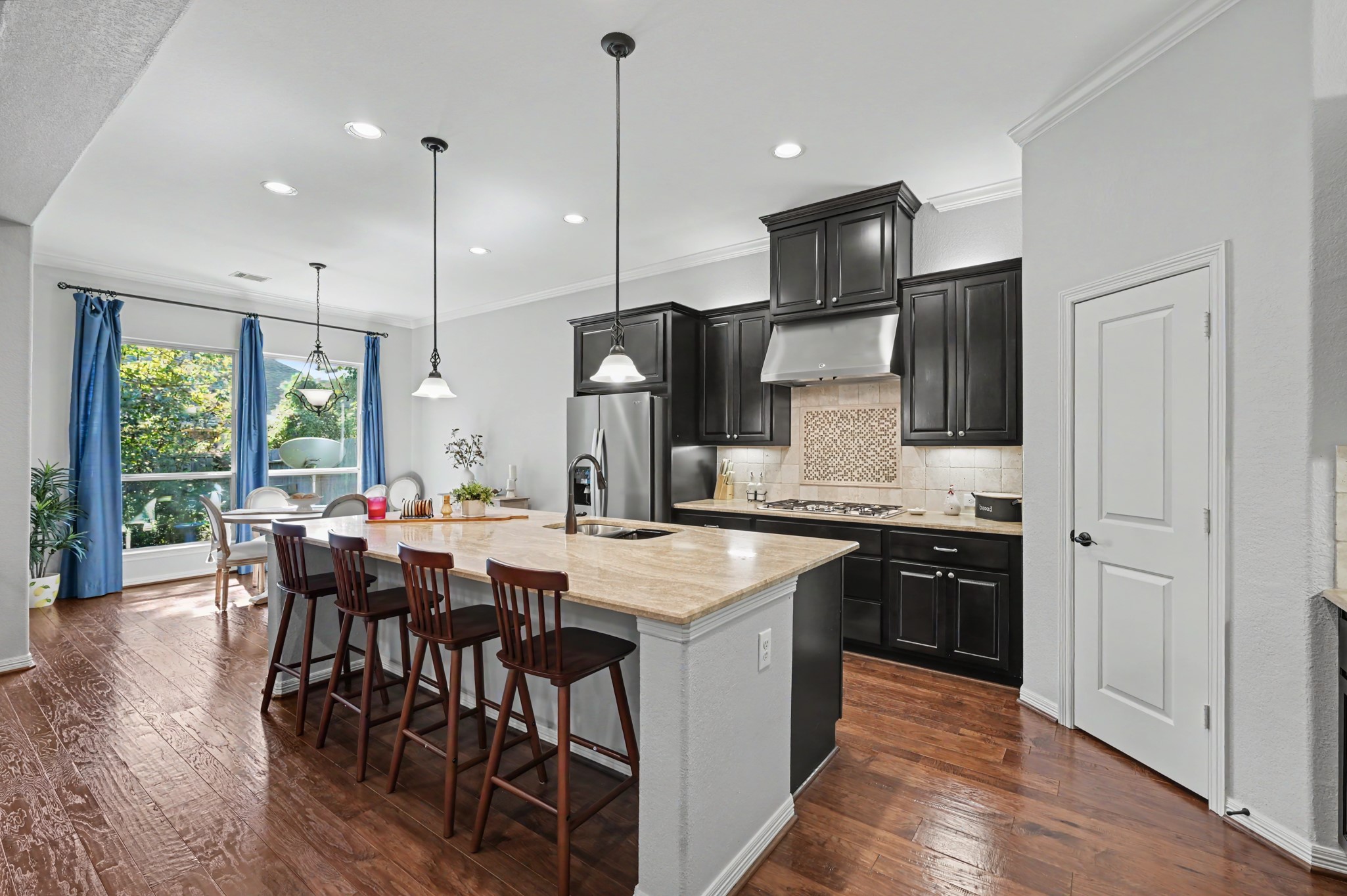 Elegant kitchen with dark cabinetry, stainless steel appliances, and a spacious island with bar seating. The open dining area is illuminated by natural light from large windows, creating a warm and inviting atmosphere.