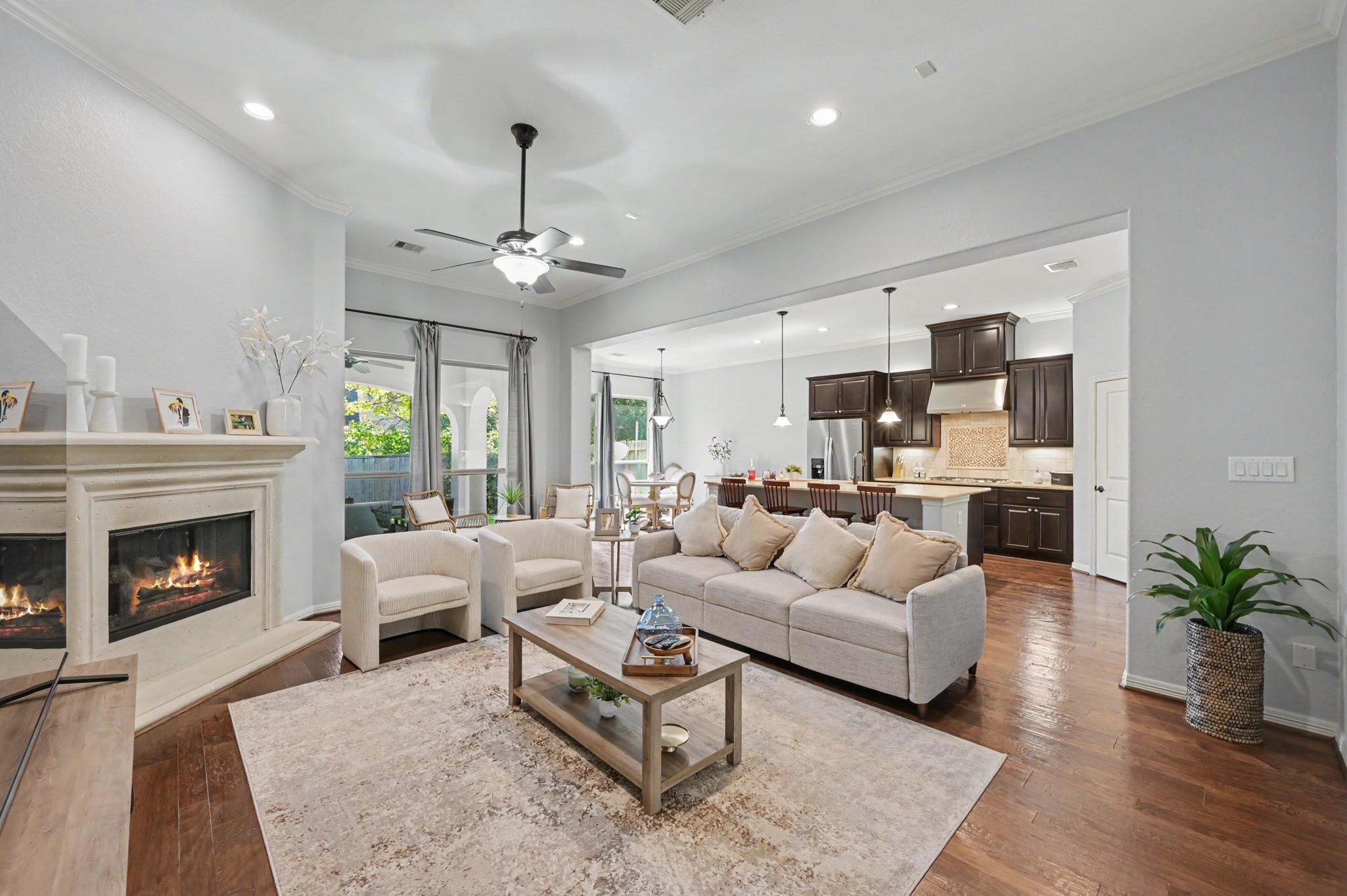 Spacious open-concept living area with a cozy fireplace, elegant hardwood floors, and seamless flow into a modern kitchen featuring dark cabinetry and a breakfast bar.