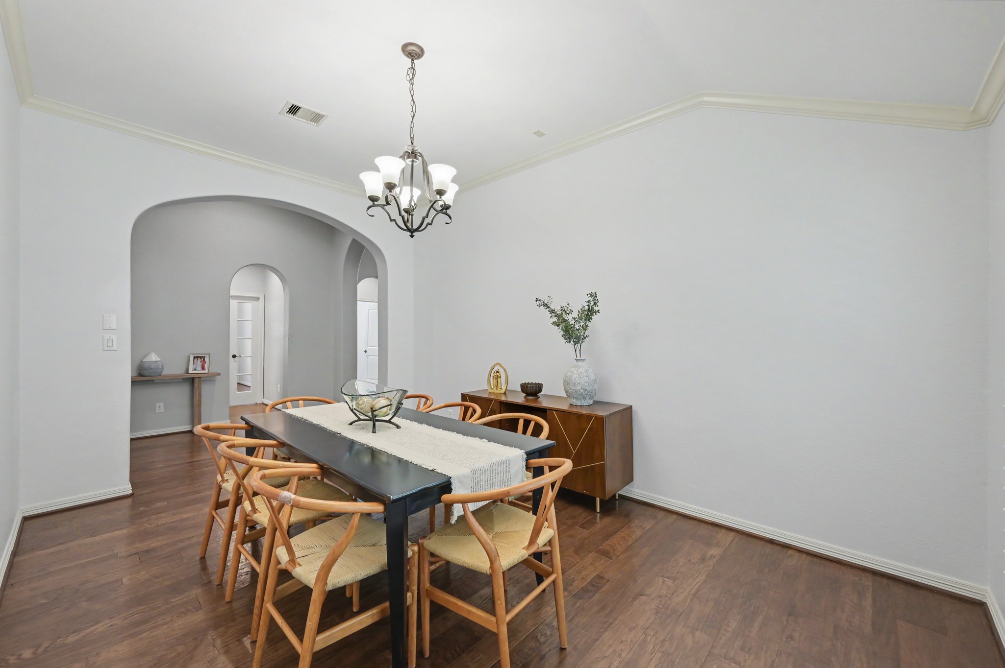 Elegant dining room with a sleek black table, surrounded by stylish wooden chairs, under a chic chandelier. Soft archways and light walls create an inviting ambiance.