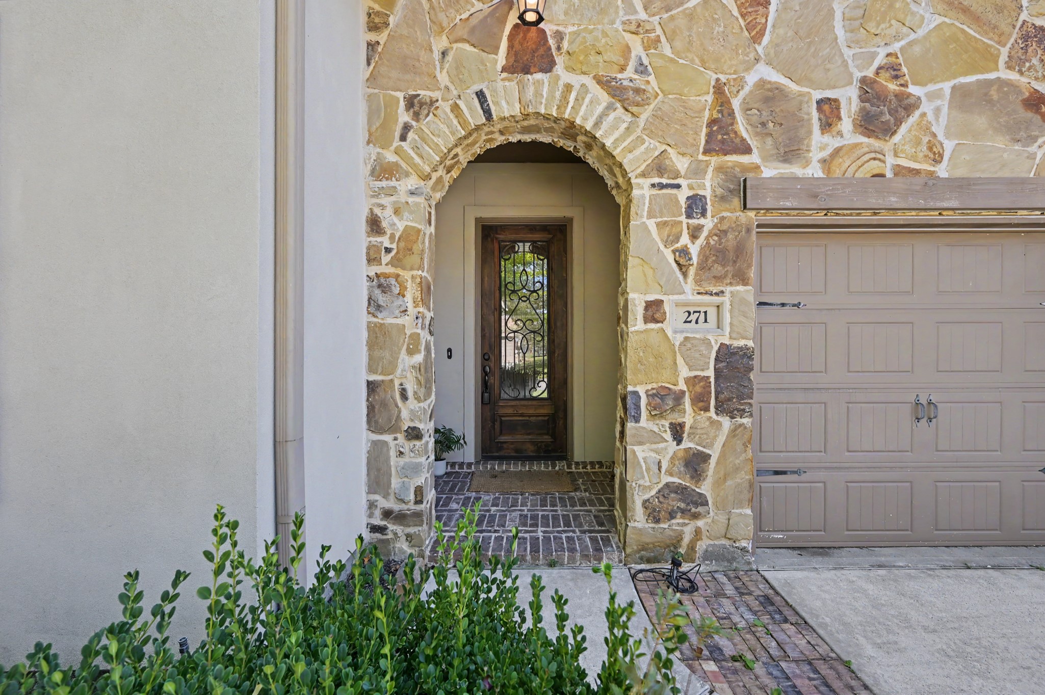 Charming stone faade with an inviting arched entryway leads to a beautifully detailed wooden front door, complemented by a stylish garage.