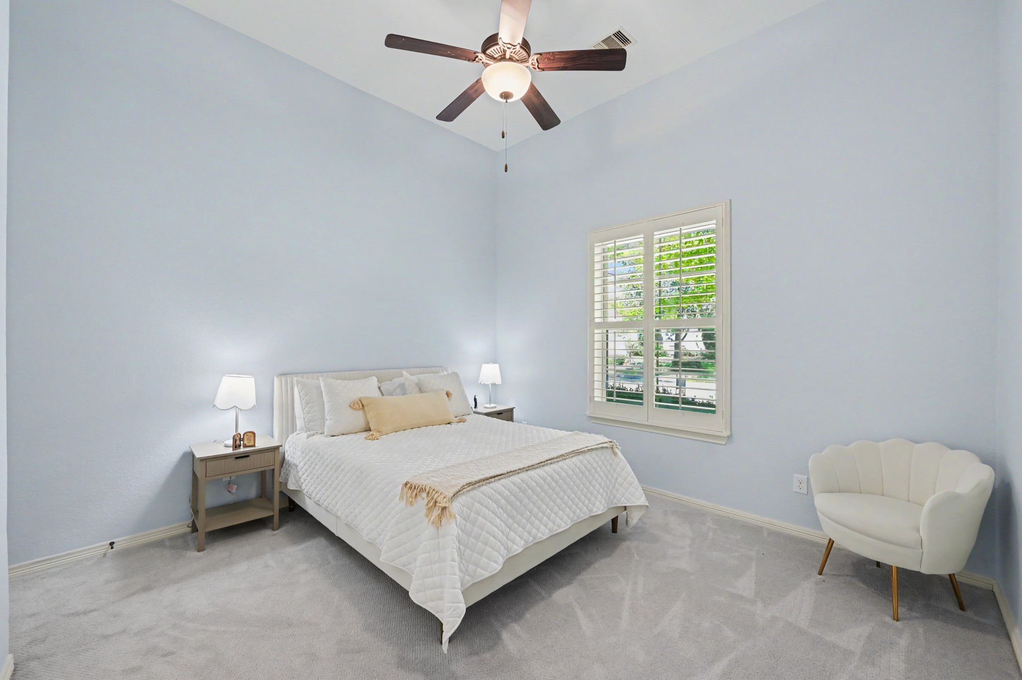 Bright and inviting bedroom with soft blue walls, plush carpeting, and a ceiling fan for comfort. The room features a cozy bed, elegant nightstands, and a charming window with plantation shutters that allow natural light to fill the space.