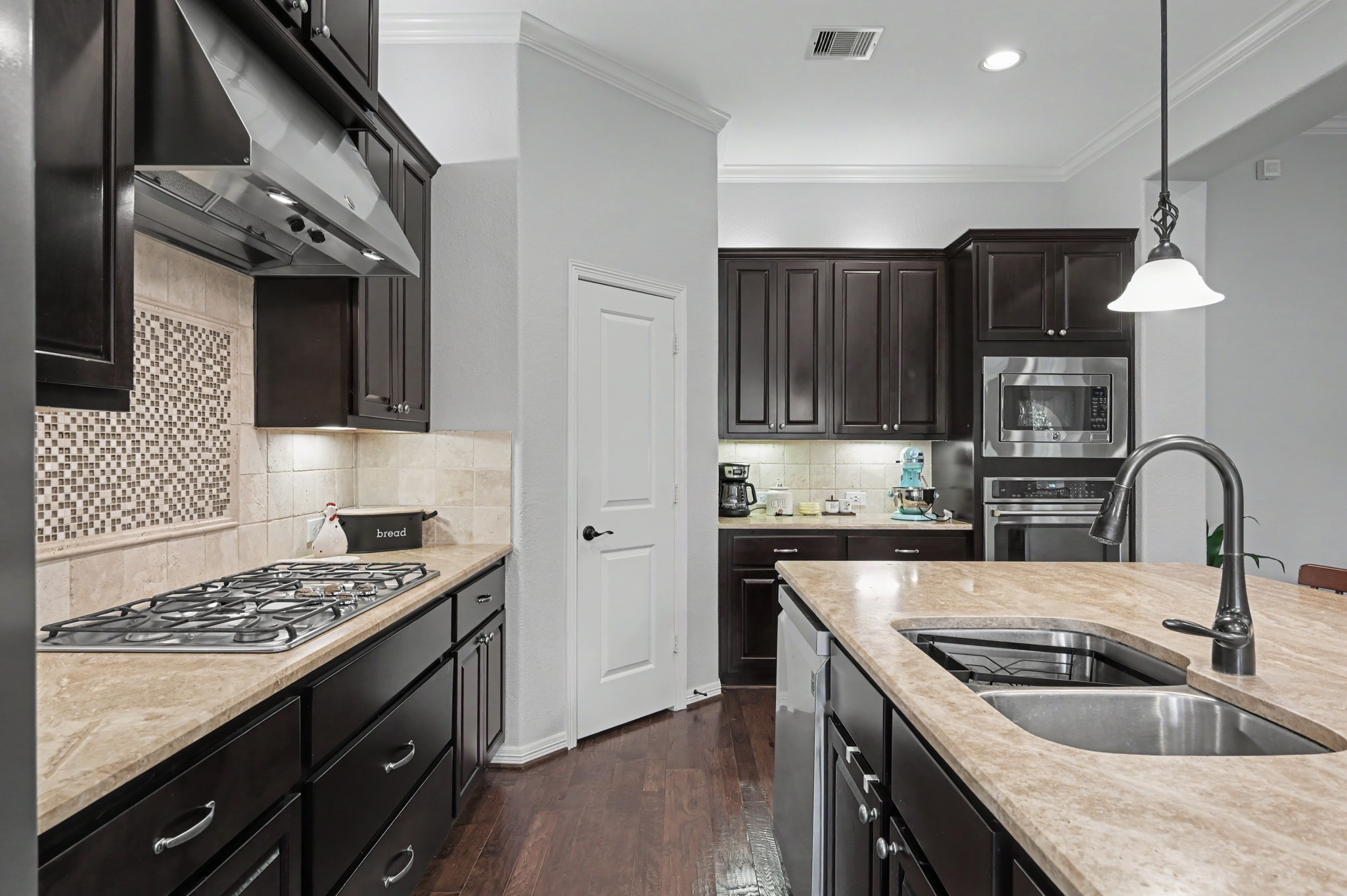 Elegant kitchen with dark wood cabinetry, granite countertops, and a central island featuring a modern sink. Includes a gas cooktop, built-in ovens, and stylish tile backsplash.