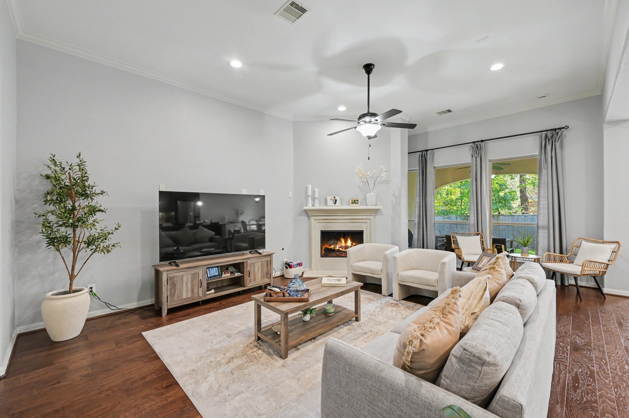 Charming living room with a cozy fireplace, elegant ceiling fan, and large windows offering natural light. Comfortable seating and stylish decor create a welcoming atmosphere.