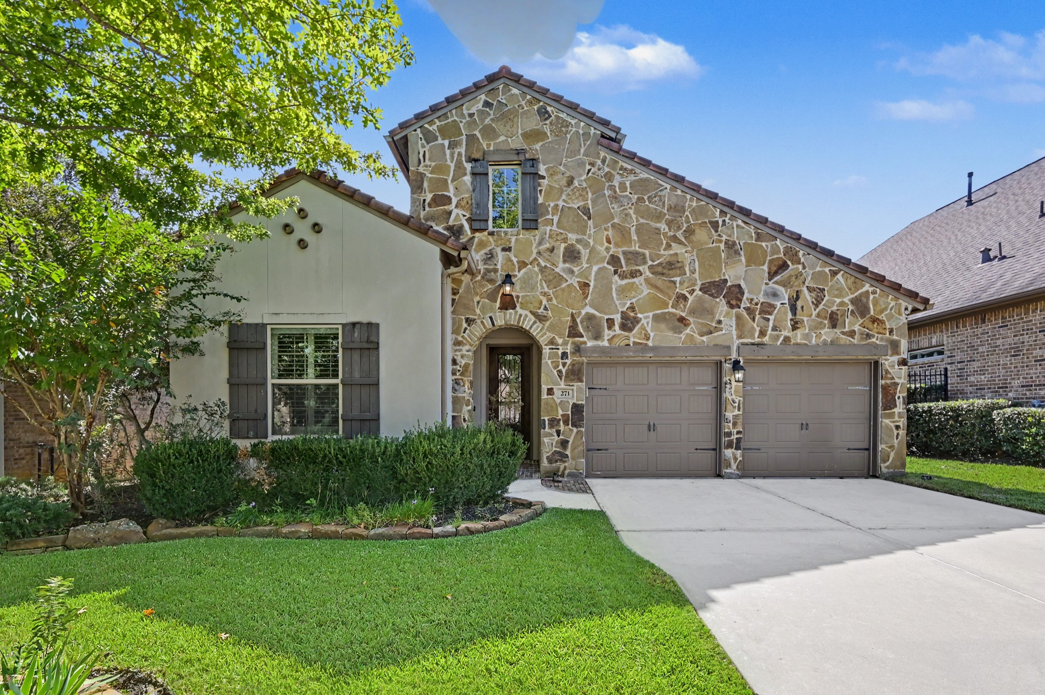 Charming home with a stunning stone facade and cozy shutters, complemented by a well-maintained lawn and lush landscaping.