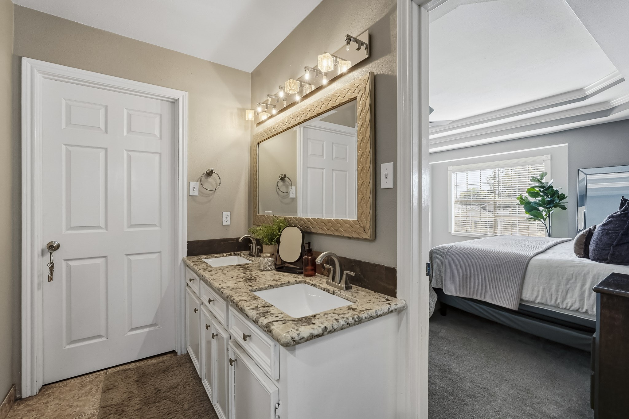Elegant primary bathroom with a dual-sink vanity, granite countertops, and a large mirror, adjacent to a cozy, well-lit bedroom.