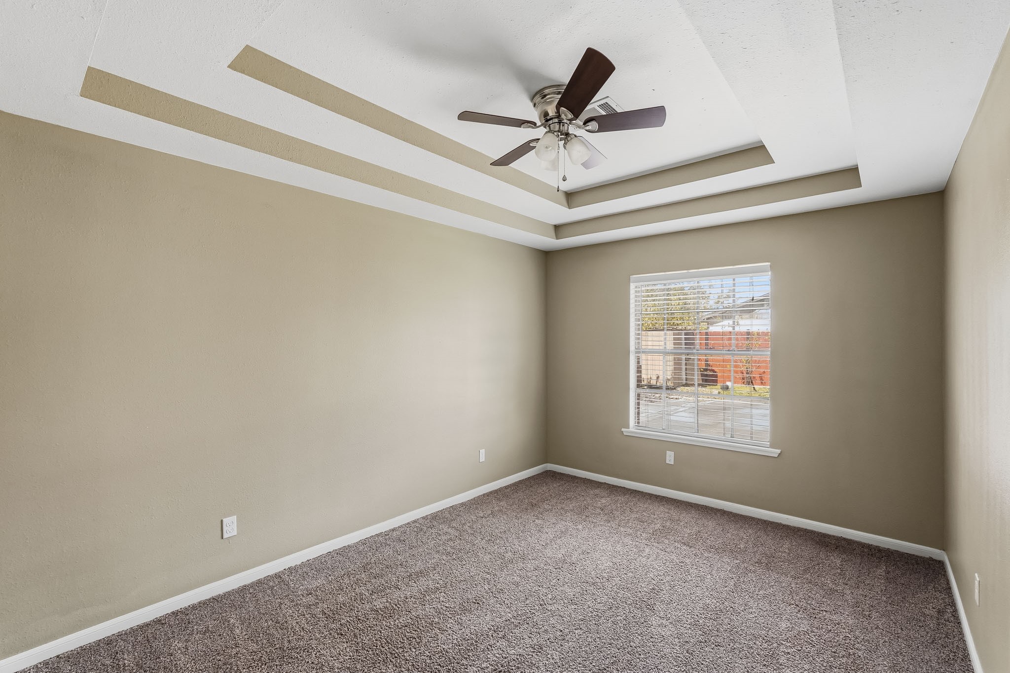 Spacious secondary bedroom with a stylish tray ceiling, ceiling fan, and soft carpeting, offering ample natural light through a large window.