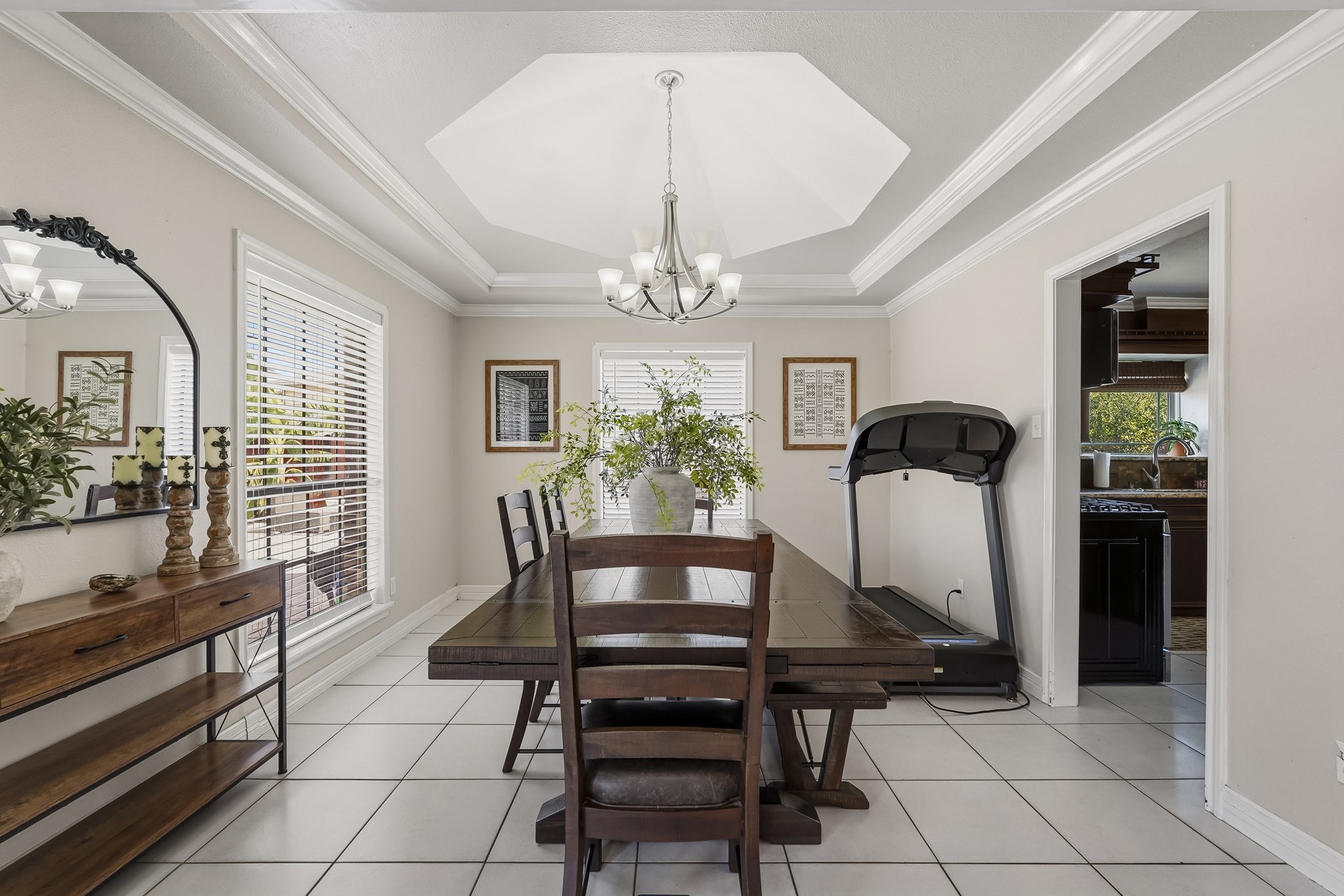 Bright dining room with elegant tray ceiling, modern chandelier, and large windows allowing natural light. Features a spacious wooden table, decorative shelving, and convenient access to the kitchen.