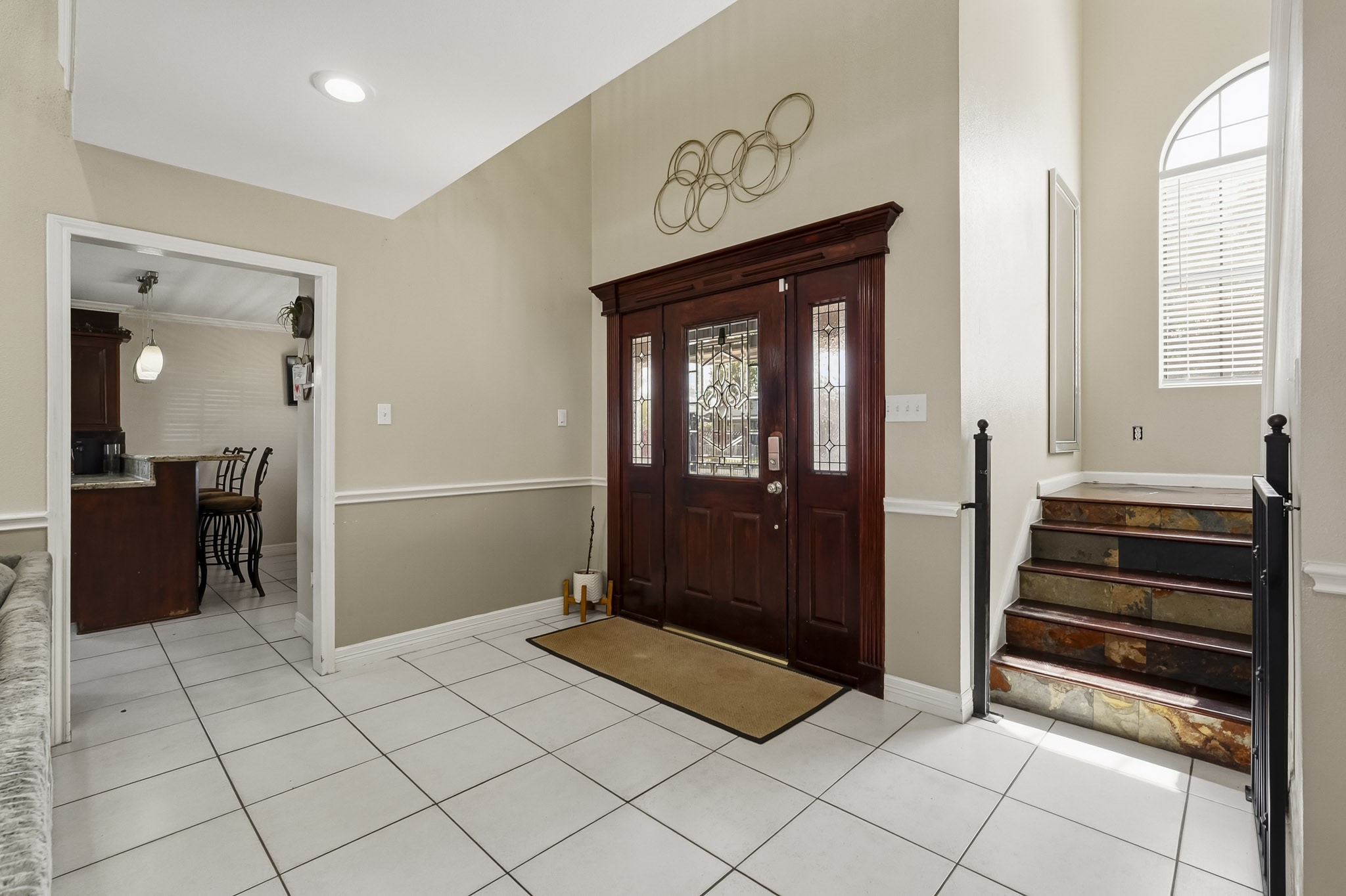 Elegant entryway with a rich wooden front door featuring decorative glass, leading to a bright, tiled foyer. An inviting staircase with wrought iron railings adds charm, while an adjacent doorway opens to a cozy kitchen area. Elegant entryway with a rich wooden front door featuring decorative glass, leading to a bright, tiled foyer. An inviting staircase with wrought iron railings adds charm, while an adjacent doorway opens to a cozy kitchen area.