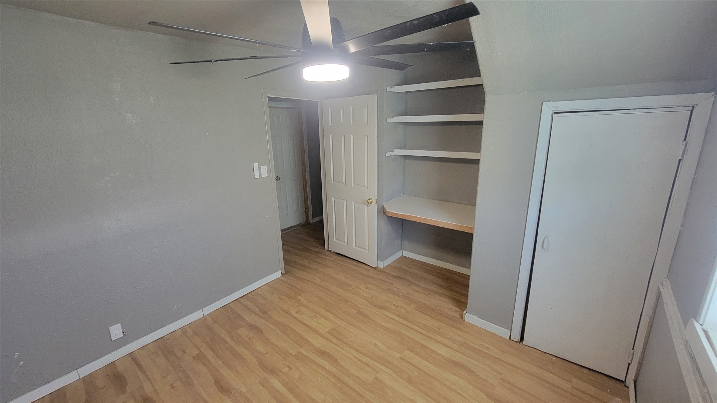 This room features laminate wood flooring, gray walls, and a modern ceiling fan. It includes built-in shelves and a small closet, providing practical storage solutions.