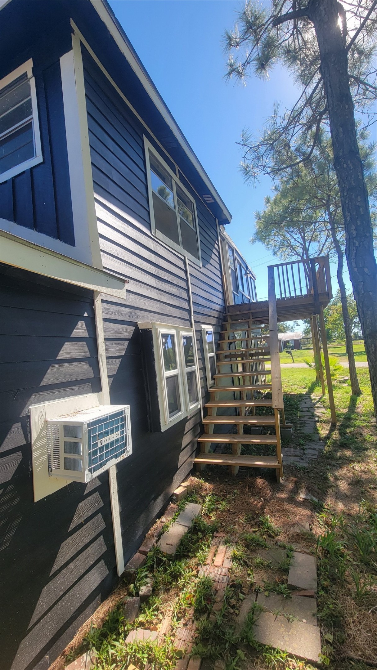 This photo shows the side of a two-story home with dark blue siding and white trim. It features an external staircase leading to an upper deck, surrounded by trees and a grassy area.