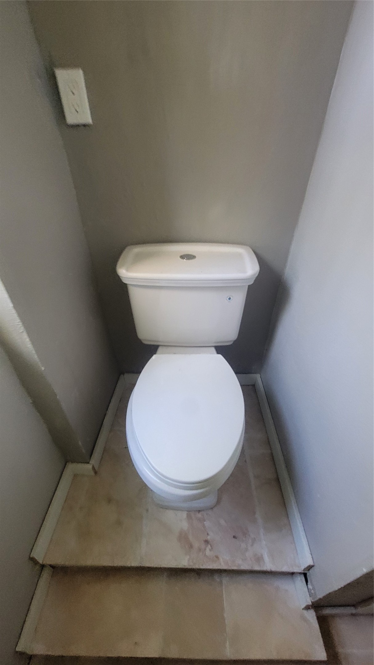 This photo shows a compact bathroom space featuring a white toilet placed on a small platform with tile flooring and gray walls.