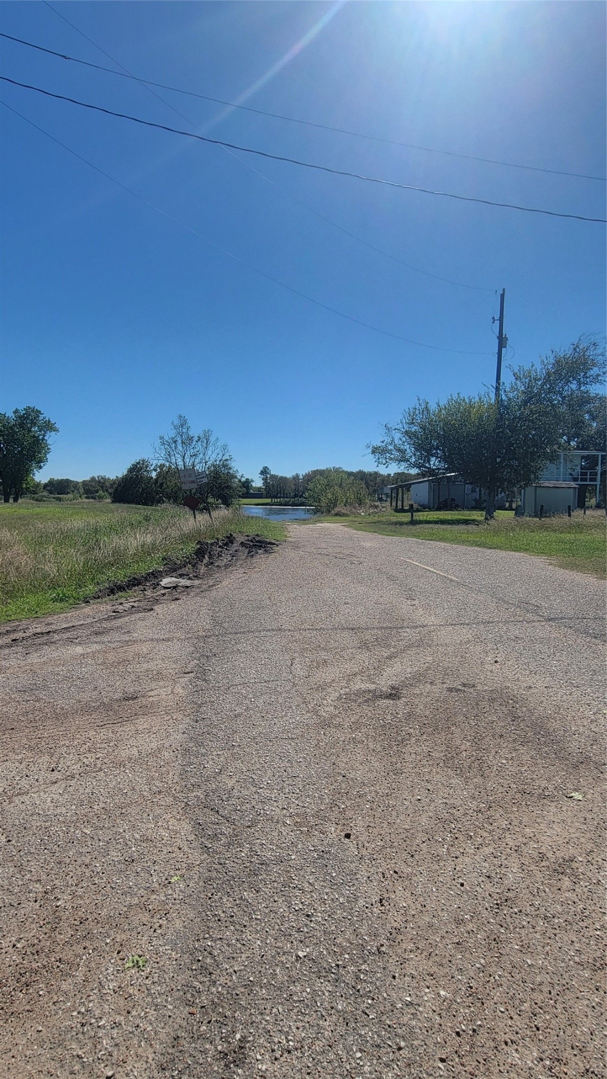 A quiet, rural road with open fields on either side and a clear blue sky. There are a few trees and a glimpse of a house to the right, offering a peaceful, country living vibe.