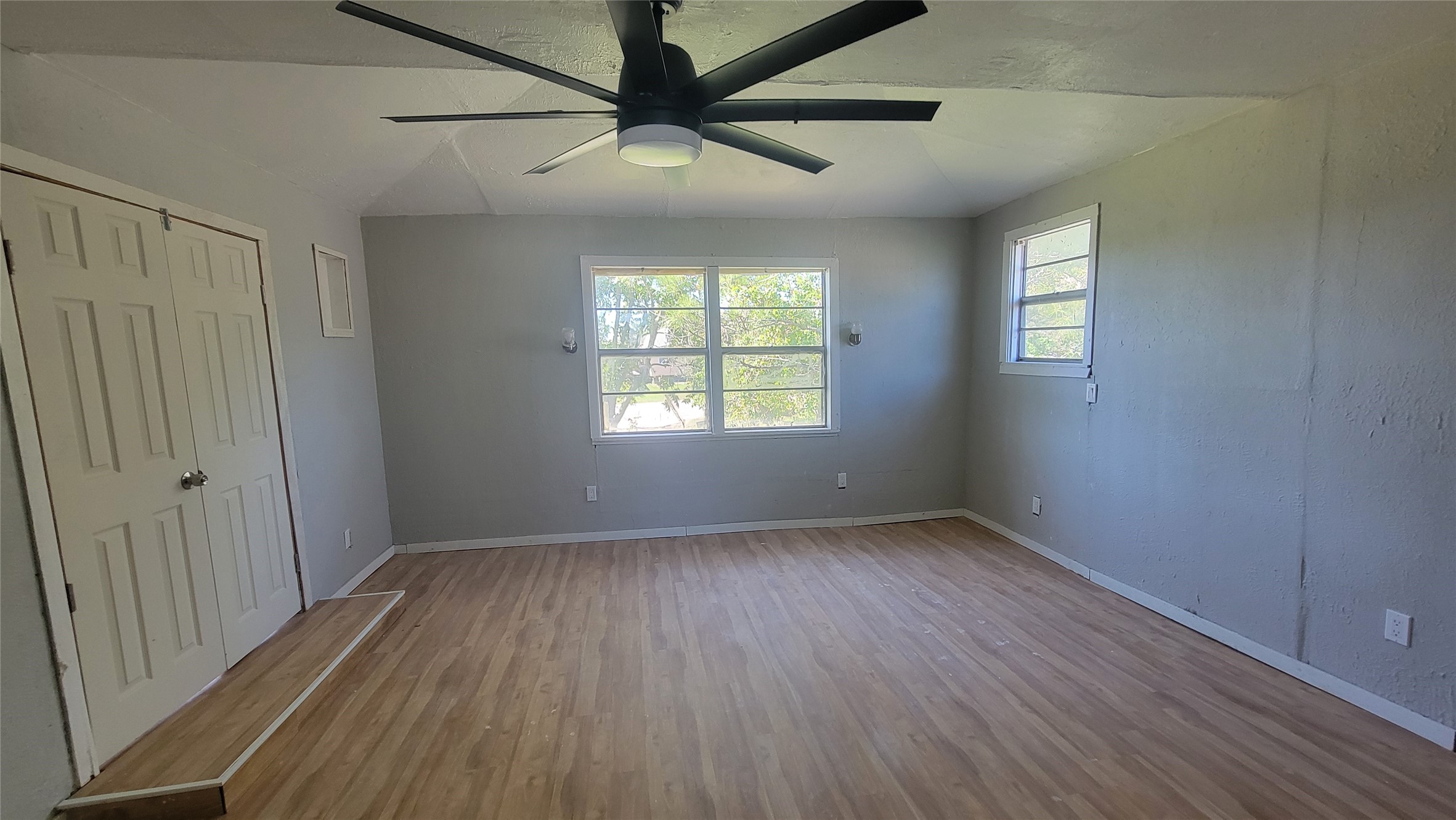 This room features a light wood floor, gray walls, a ceiling fan, and ample natural light from two windows. There is also a double-door closet for storage.