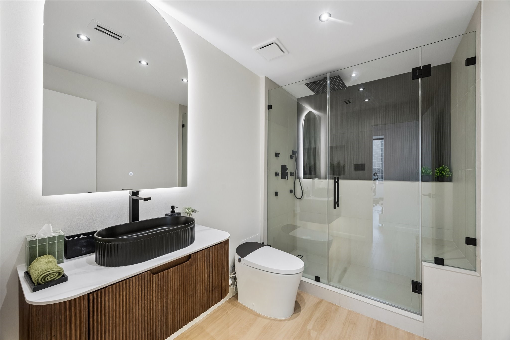 Floating fluted vanity and illuminated mirror complement a glass enclosed shower with a bench.