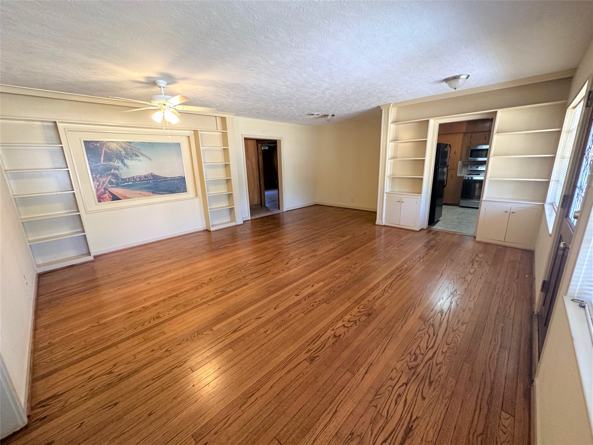 Another view of large living room with tons of built-ins, ceiling fan and beautiful hardwood floors. Doorway in middle leads to primary suite hallway and additional living area/den. Doorway on right leads to kitchen. Another view of large living room with tons of built-ins, ceiling fan and beautiful hardwood floors. Doorway in middle leads to primary suite hallway and additional living area/den. Doorway on right leads to kitchen.