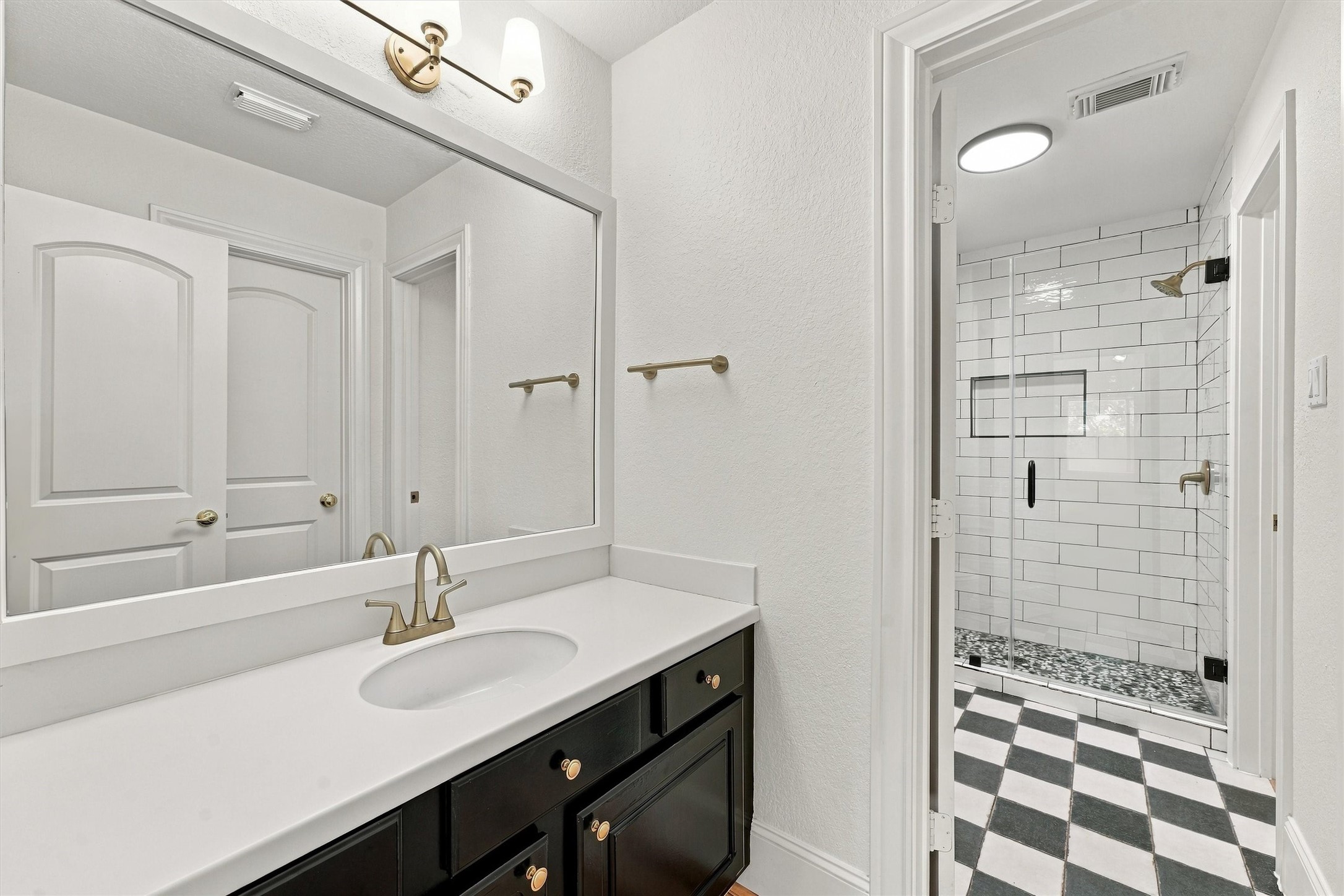 Second floor - Third Bathroom with black vanity, large framed mirror, brass fixtures, checkerboard tile floor, and a walk-in shower with white subway tile, niche, and glass door.