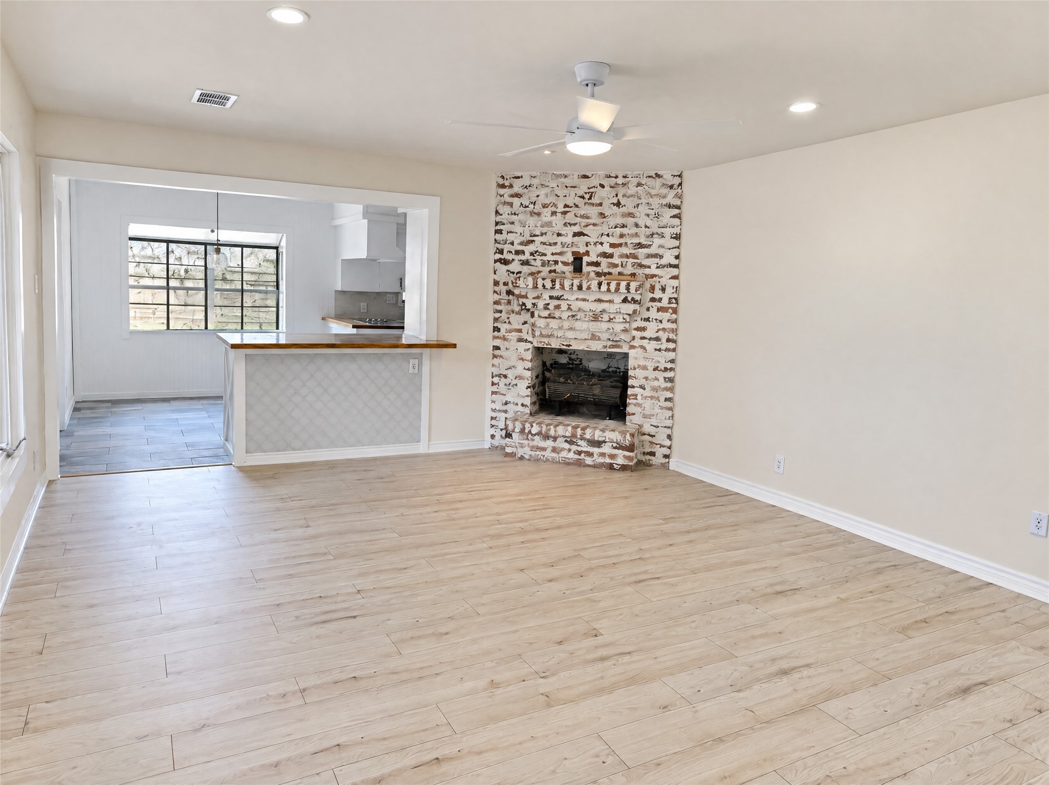 Flowing effortlessly toward the kitchen, the primary living space showcases a whitewashed brick fireplace as its centerpiece. Recessed lighting, a modern ceiling fan, and direct sightlines to the breakfast bar create a warm, connected atmosphere for gathering with family and friends.
