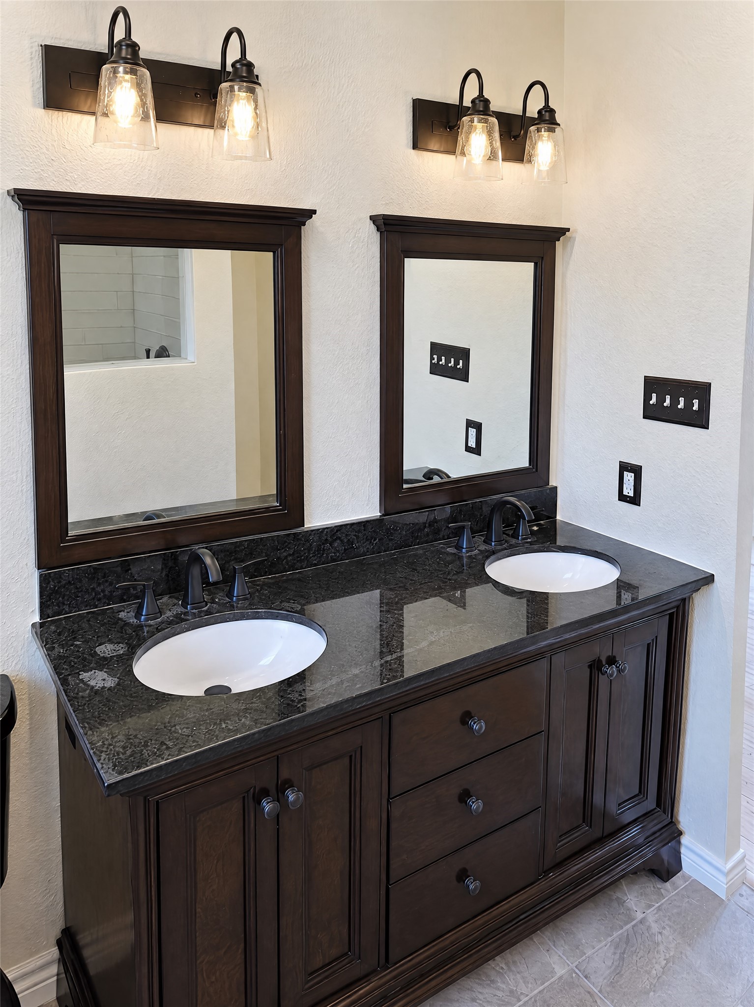 Step into the en-suite bathroom showcasing an elegant dark wood double vanity topped with polished granite, matching framed mirrors, oil-rubbed bronze fixtures, and warm designer lighting.
