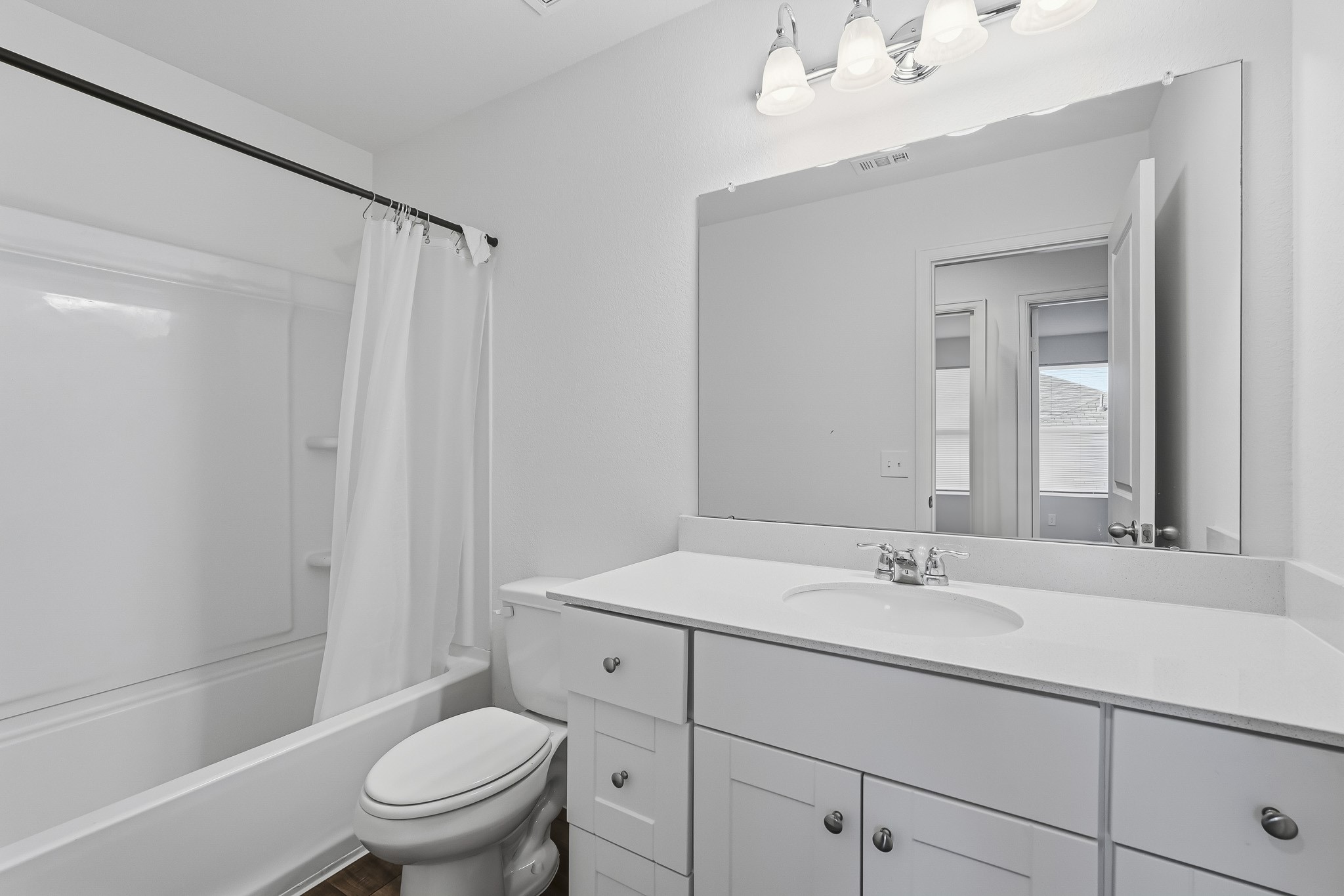 This bright and modern full bathroom features a clean, refreshing design with crisp white walls and a timeless neutral color palette. A spacious vanity offers generous countertop space, cabinetry, and ample storage, complemented by a large wall-mounted mirror and elegant four-light fixture. The shower and bathtub combination features a white surround with built-in shelving for added convenience.