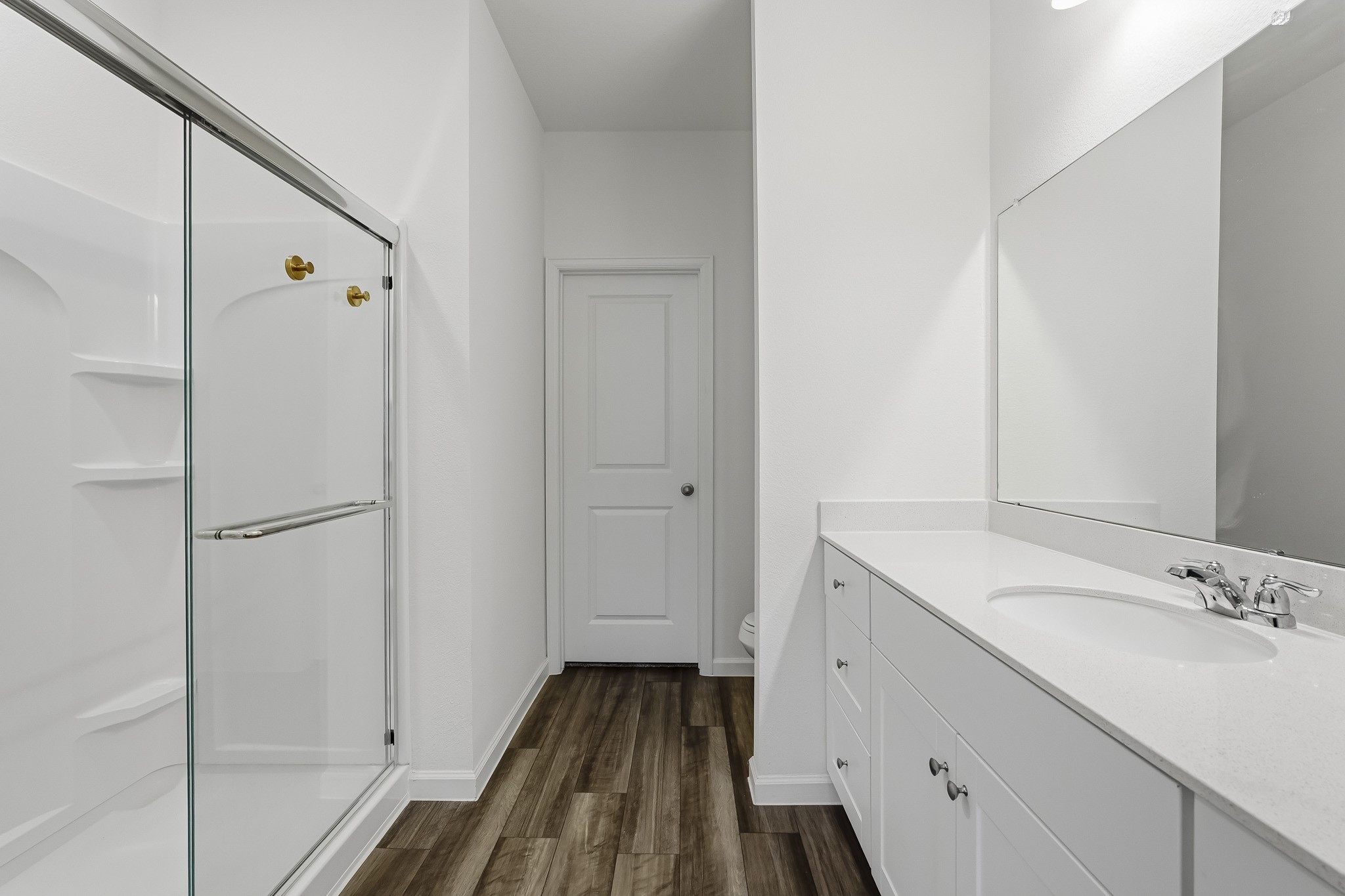 This bright and spacious primary ensuite bathroom features a clean, modern design with crisp white walls, rich wood-look flooring, and elegant finishes throughout. At the back of the bathroom, a separate door leads to a walk-in closet, providing convenient storage and easy access to wardrobe essentials.