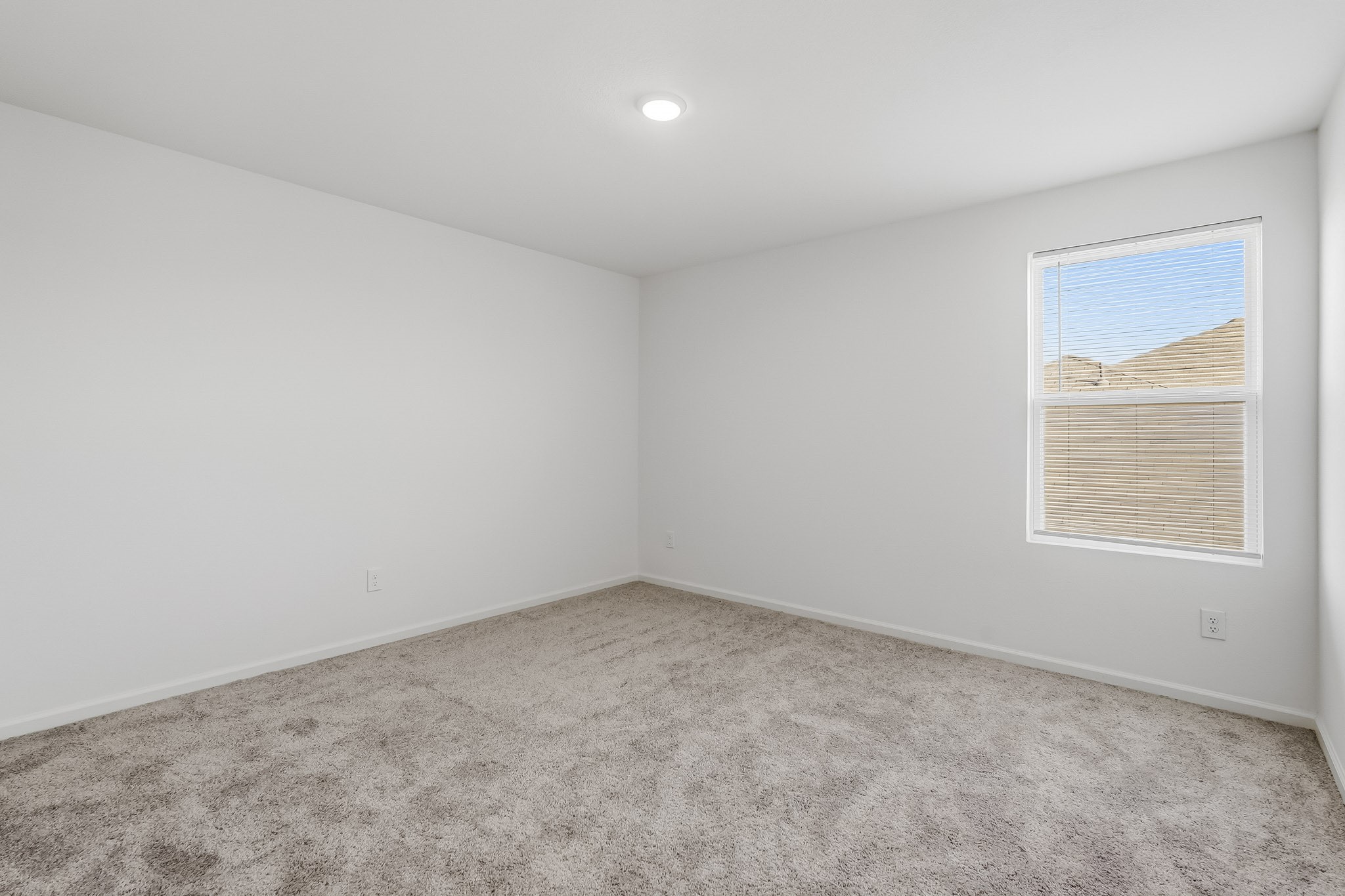 This bright and inviting bedroom features a spacious layout with crisp white walls, plush neutral carpeting, and a clean, contemporary design. A generously sized window with adjustable blinds allows natural light to fill the space, creating a fresh and airy atmosphere.